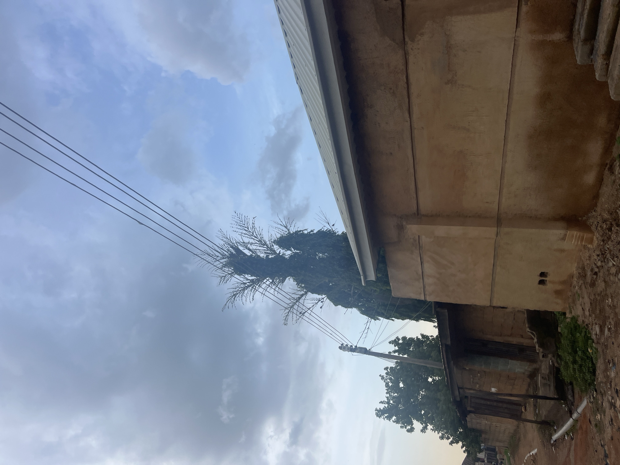 The image shows a residential street scene in Jos, Nigeria, under a cloudy sky. In the foreground, a weathered, light brown building with a corrugated metal roof occupies the right side of the frame. Its wall has a horizontal line running across it and two square openings near the bottom. To the left of this building, there is a smaller, older structure with a wooden awning and what appears to be a dirt or mud brick wall. This structure has two doorways, one of which is a wooden door. In front of these buildings, a dirt road or path is visible, with some loose stones and debris on the ground. A white pipe runs along the edge of the road. Several trees are present in the background, including a tall, slender tree with sparse foliage, and other trees with denser canopies. Power lines are strung across the upper part of the image, supported by a utility pole on the left. The overall impression is one of a modest, somewhat rural or older urban area.