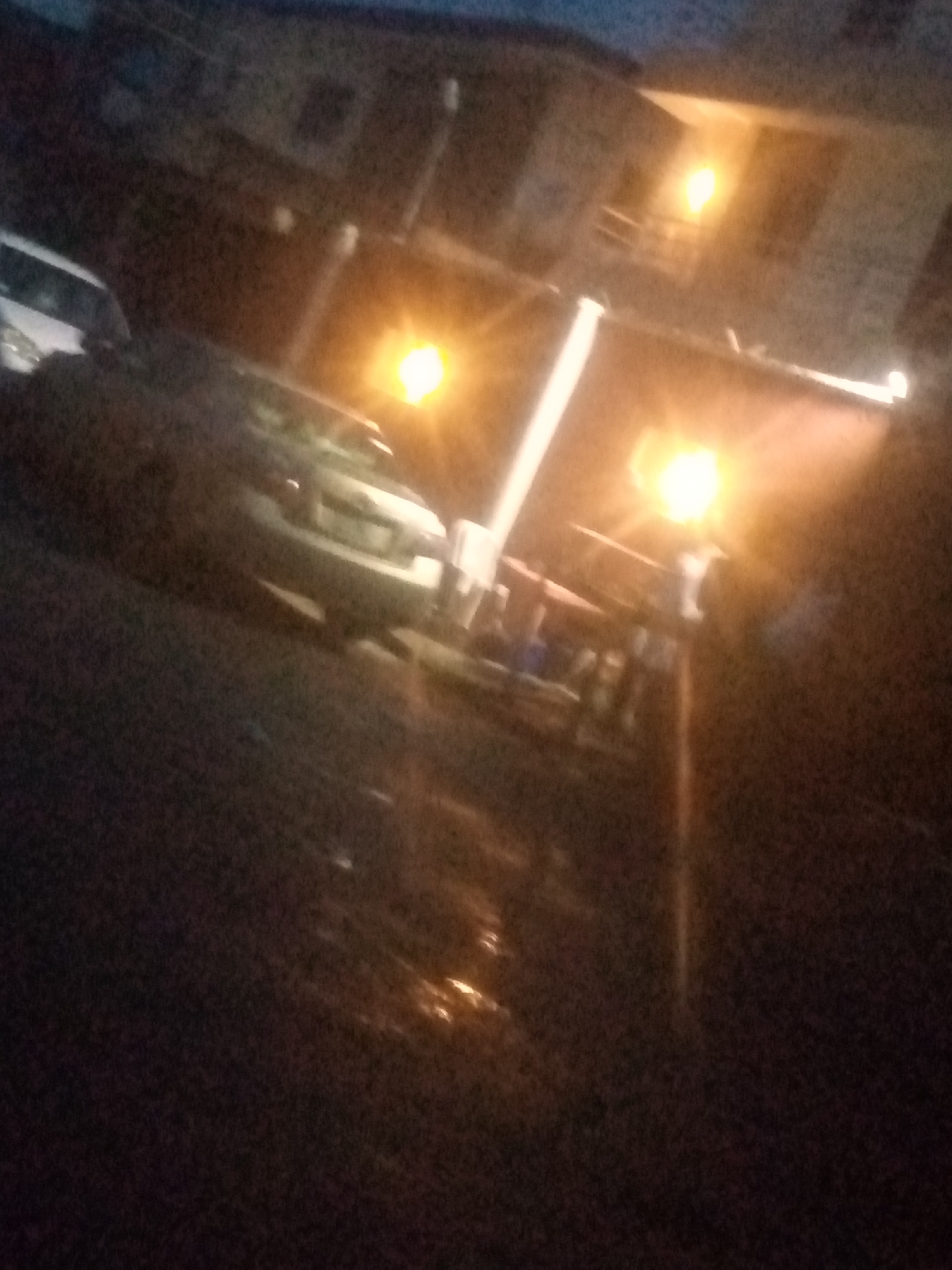 The image presents a severely blurry, dark, and grainy nighttime street scene in Shangisha, Nigeria, characterized by poor focus and low-light conditions. Artificial illumination from multiple bright, warm-toned light sources pierces the darkness, creating lens flares and strong reflections.
On the far left, a portion of a white car is visible, angled towards the right. Further into the mid-ground, a light-colored sedan is positioned on a dark, reflective surface, which appears wet, possibly from rain. In the upper right background, a building with an illuminated window or balcony is discernible.
The mid-right section of the frame features an outdoor commercial establishment or covered area, marked by prominent vertical supports and overhead structures. This area is brightly lit by at least two distinct artificial light fixtures. Beneath these lights, indistinct, shadowy figures of individuals are visible, suggesting people gathered around tables, possibly engaged in dining or socializing. The ground in the foreground is dark and textured, showing patches of light reflection. The scene conveys an urban or semi-urban environment at night, with vehicular presence and human activity concentrated around the illuminated outdoor space.