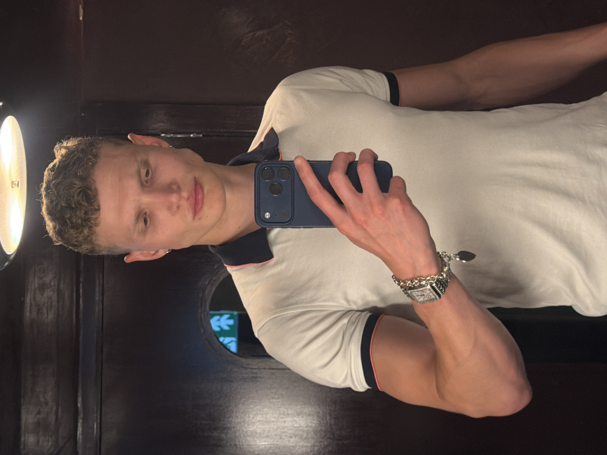 A young man with curly blonde hair, an earring, and a light complexion stands in front of a dark wood paneled wall, holding a dark blue smartphone in his right hand, taking a selfie. He is wearing a white polo shirt with a dark blue collar and orange trim, and a silver bracelet with a heart-shaped charm on his left wrist. The background is dimly lit, with a circular illuminated sign visible through an archway behind him, displaying green directional arrows. The overall impression is of a candid mirror selfie taken in an indoor, possibly urban, setting such as a restroom or a dimly lit corridor within a building. The location is not specified as London, United Kingdom.