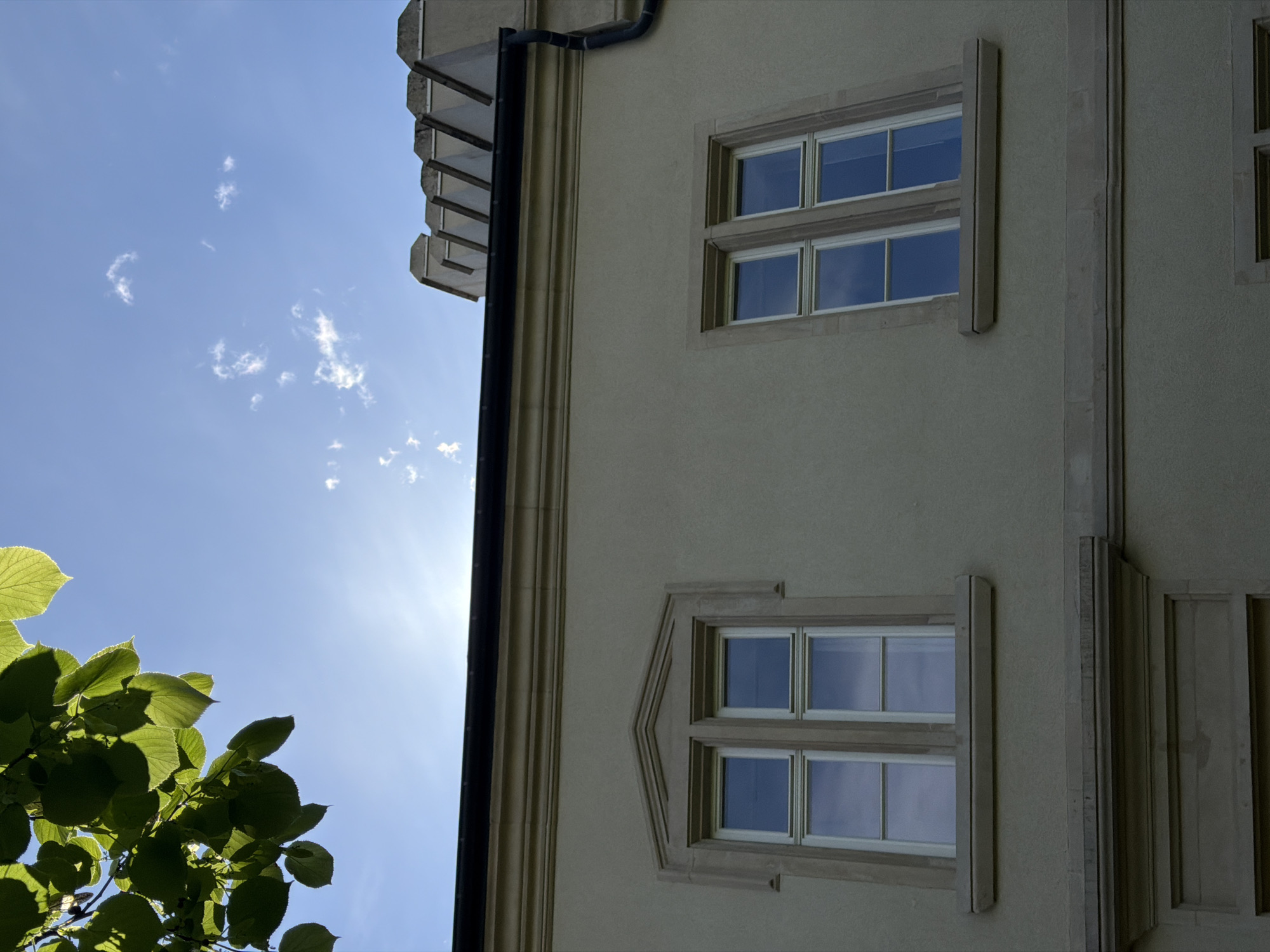 The image features a low-angle view of a building in Esch-sur-Alzette, Luxembourg, with a blue sky and some foliage in the foreground. The building is light-colored with architectural details such as window frames and vertical lines. Two windows with four panes each are visible. The sky is bright with scattered clouds. A section of green leaves and branches is visible in the lower-left corner. A black drainpipe runs vertically along the building's corner.