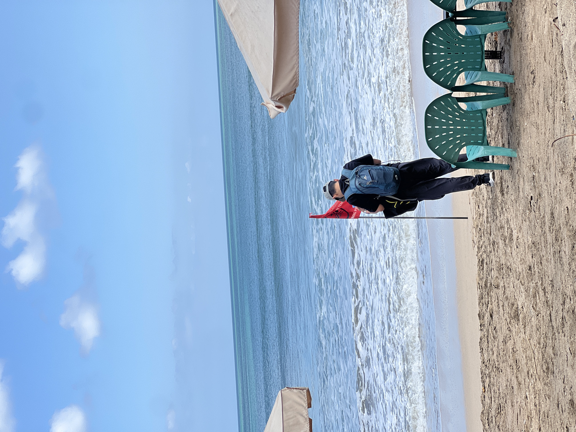 In Kuta, Indonesia, a person wearing a black t-shirt, black pants, a grey baseball cap, and a blue backpack stands on a sandy beach facing the ocean. The person is positioned near a red flag planted in the sand. Behind the person, three green plastic chairs are arranged in a row. The ocean is visible with waves breaking on the shore, and the sky is blue with scattered white clouds. A tan beach umbrella is partially visible on the right side of the frame.