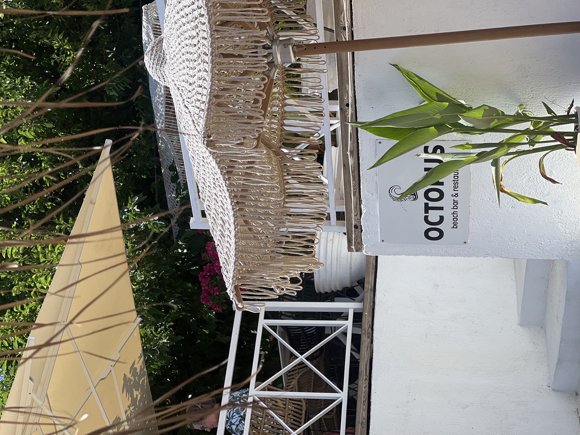 The image depicts an outdoor seating area at a beach bar and restaurant in Achladias, Greece. In the foreground, a white sign with the text "OCTOPUS beach bar & restau" is visible, accompanied by a logo featuring an octopus. A potted plant with large green leaves stands to the right of the sign. Behind the sign, a macrame umbrella with fringe detail is prominently displayed. In the midground, a white railing separates the seating area from a lower level. Wicker chairs and tables are visible behind the railing. A person is seated at one of the tables, partially obscured. Above the railing, a large beige umbrella provides shade. Lush green foliage and bright pink flowers are visible in the background, suggesting a Mediterranean setting. The overall scene conveys a relaxed, outdoor dining atmosphere.
