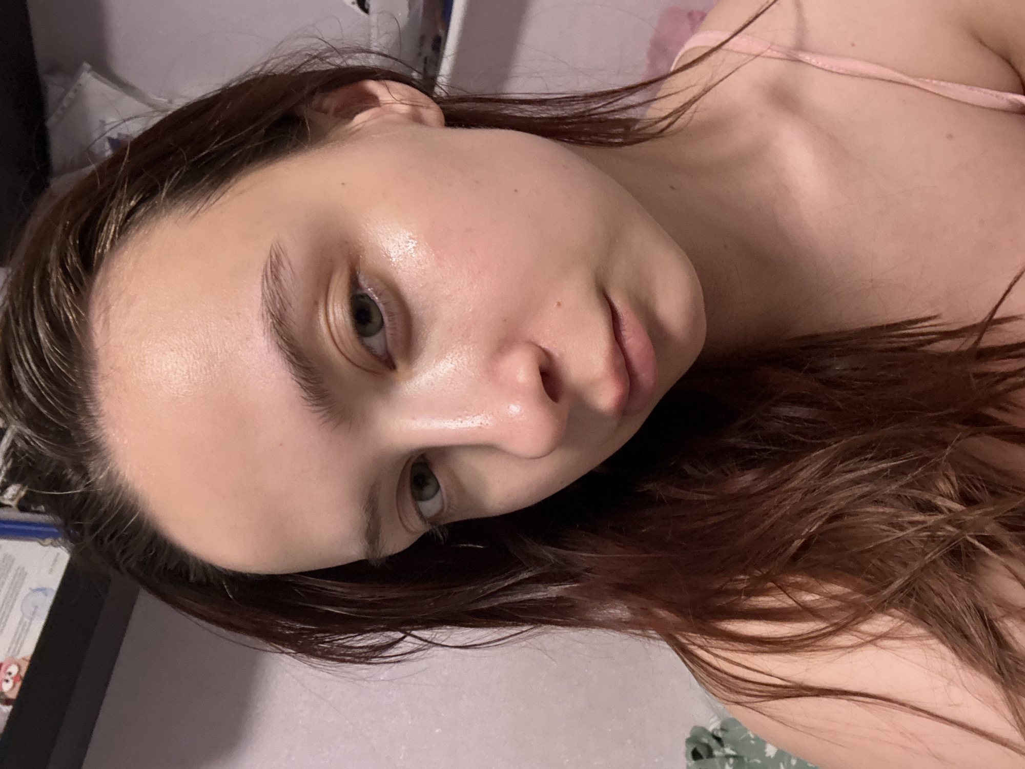 A close-up, tilted, indoor photograph depicts a young woman with light skin, dark brown hair, and light-colored eyes. Her hair appears slightly wet and is draped around her face and shoulder. She wears a light pink tank top. The background includes portions of a bed with pink and white bedding, as well as a dark object that is likely a headboard or other furniture. The photo does not provide information to determine the location in the city of Прикамский, Russia.