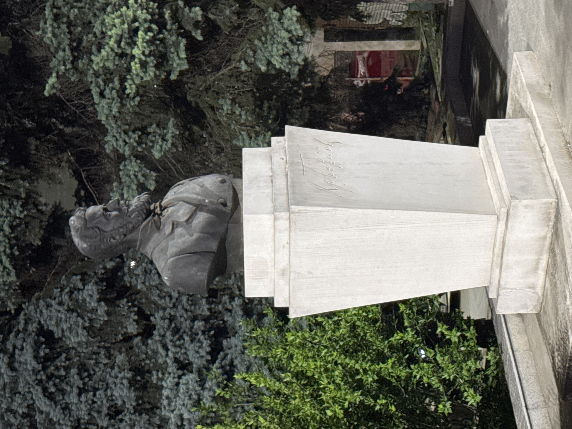 The image shows a statue of Petko Rachov Slaveykov in Veliko Tarnovo, Bulgaria. The bust of the statue is on a stepped stone pedestal with Slaveykov's name inscribed on it. The statue is positioned outdoors. Vegetation surrounds the statue, which includes bushes and trees. A building, possibly residential, can be seen in the background. The time of day is likely dusk or night. The statue is the subject of the photograph.