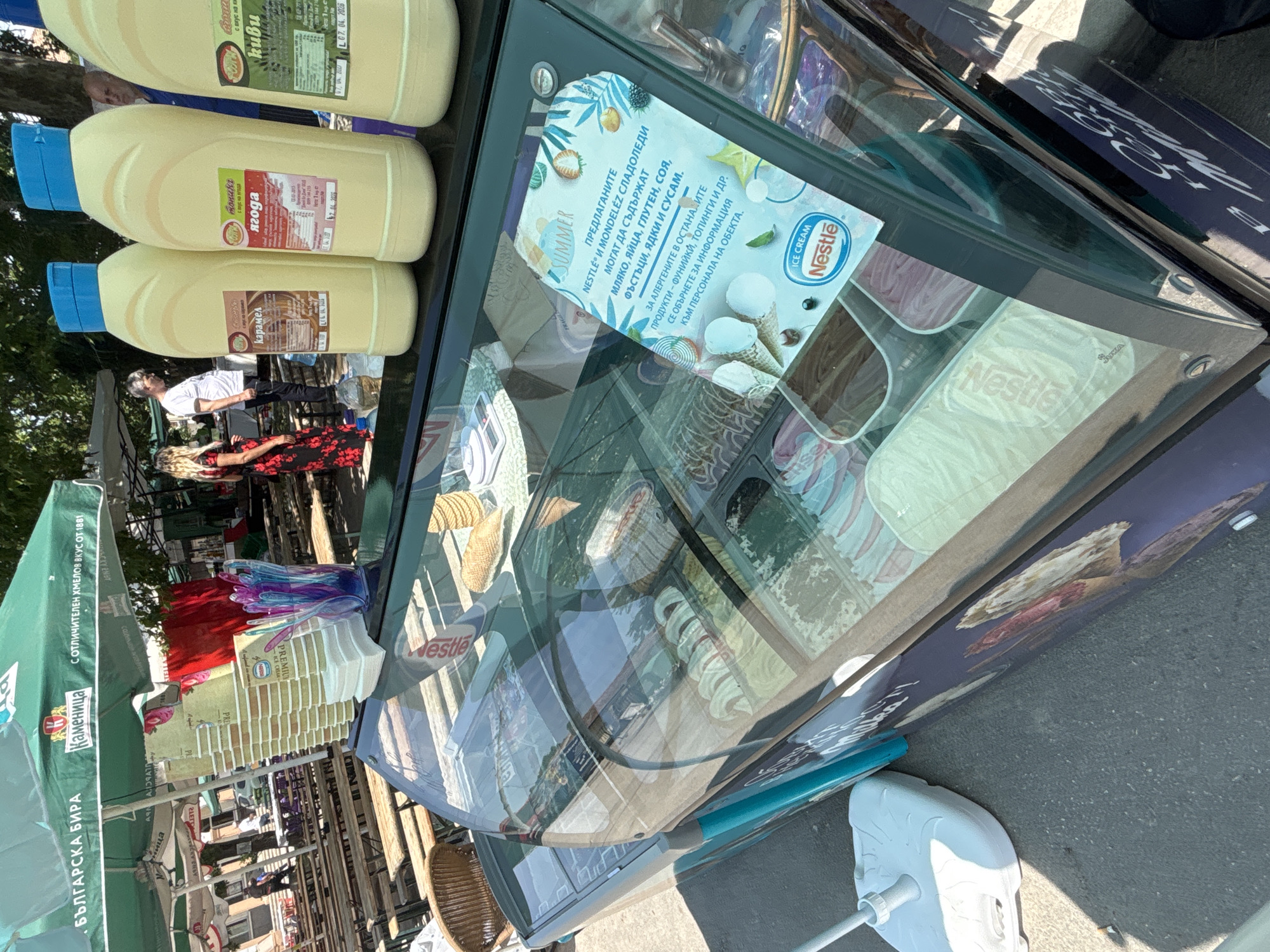 This image captures an outdoor ice cream stand operating in the city of Kostievo, Bulgaria. The stand features a glass-fronted display case showcasing various flavors of ice cream, some with visible Nestlé branding. On top of the display case, three large yellow plastic bottles with blue caps are arranged, labeled in Bulgarian as "карамел" (caramel), "ягода" (strawberry), and "киви" (kiwi), indicating ice cream toppings. A stack of Nestlé-branded disposable cups, a pile of napkins, and a clear container holding colorful plastic spoons or spatulas are also present on the counter. An allergy information sticker on the display case's glass front, in Bulgarian, advises that Nestlé and Mondelez ice creams may contain milk, eggs, gluten, peanuts, nuts, soy, and sesame, and directs customers to ask staff for information on other products.
In the background, a large green umbrella with "Каменица" (Kamenitza) and "БЪЛГАРСКА БИРА" (BULGARIAN BEER) branding provides shade over multiple rows of wooden picnic-style tables and benches. Several individuals are visible in the scene: a woman in a red floral dress stands to the left of the ice cream stand, facing a man in a white t-shirt and dark pants who appears to be behind the stand. Another man in a blue shirt is partially visible on the far right. The ground consists of asphalt or pavement, and the scene is brightly lit by daylight, casting distinct shadows. A white umbrella base is visible on the ground to the left of the ice cream stand.