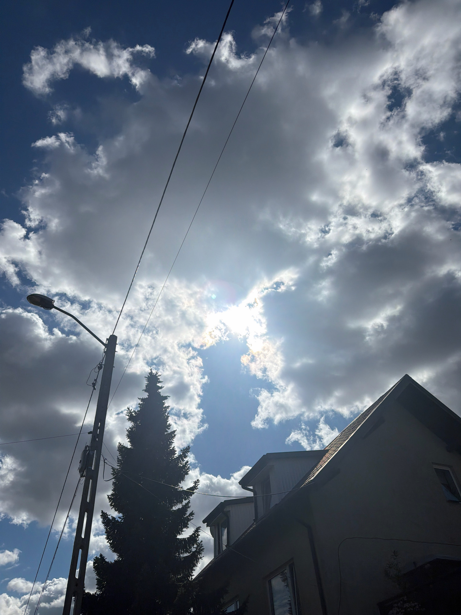 This is an outdoor, low-angle, full-shot photograph of a cloudy sky with sunlight peeking through the clouds in Szczecin, Poland. In the foreground and to the left, a tall, grey concrete lamppost with a black lamp fixture on a curved arm is visible, with multiple wires attached to it. A tall, dark green, coniferous tree stands to the right of the lamppost. In the lower right quadrant of the image, the roof and upper portion of a multi-story building with visible windows is silhouetted against the bright sky. The sky itself is a mix of dark and light grey clouds, with patches of blue sky visible. Sunlight is breaking through the clouds in the upper right portion of the image, creating lens flare.