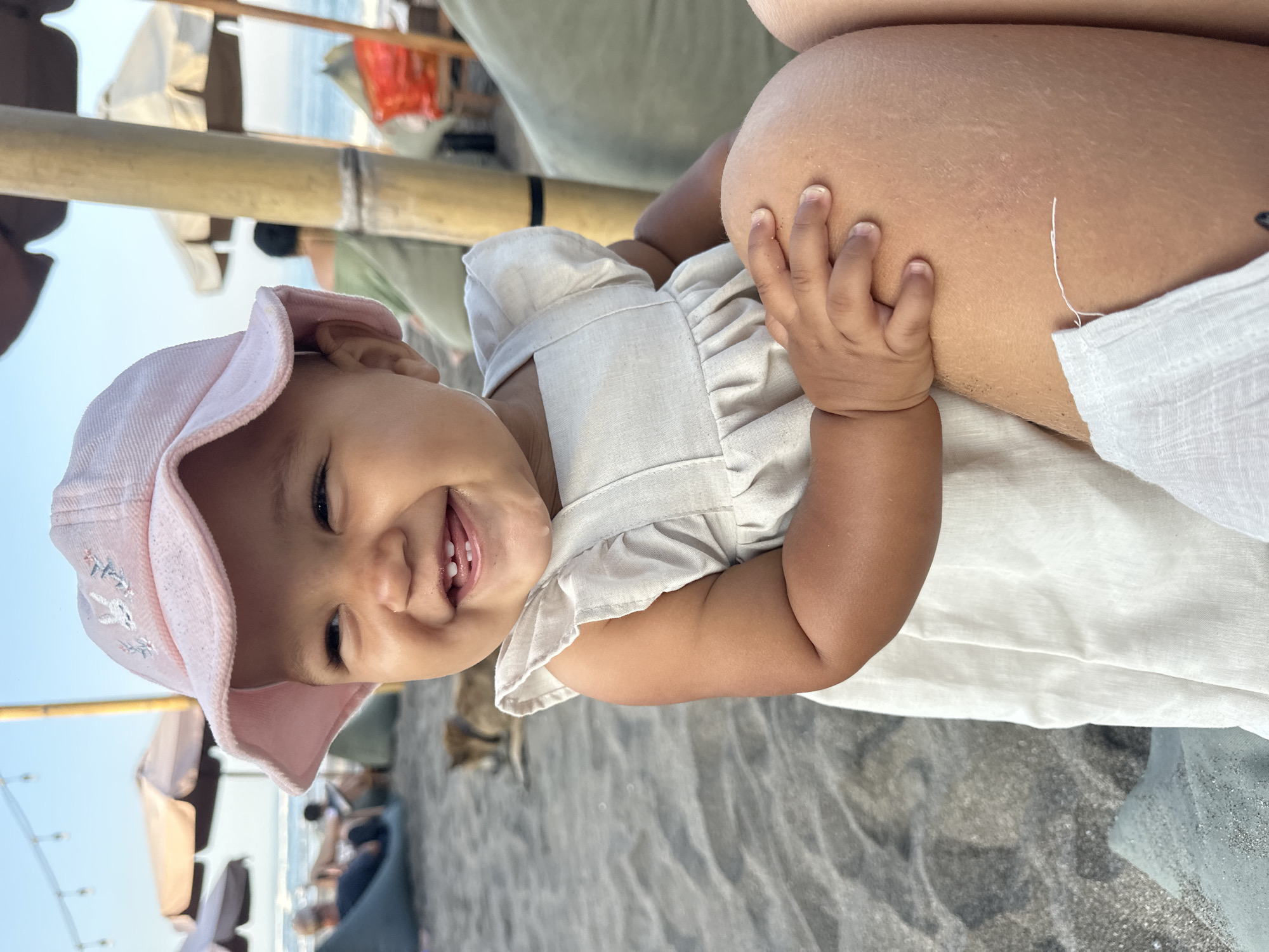 A smiling baby girl wearing a pink bucket hat and a light-colored dress sits on a sandy beach in North Kuta, Indonesia. She is looking to her left, with her eyes wide and her mouth open in a happy expression, revealing two tiny teeth. Her small hand is resting on a person's knee. In the background, beach umbrellas, lounge chairs, and people relaxing can be seen, as well as a dog lying down on the sand. The ocean and sky are visible in the distance. The overall atmosphere appears to be relaxed and joyful, indicative of a beach outing.