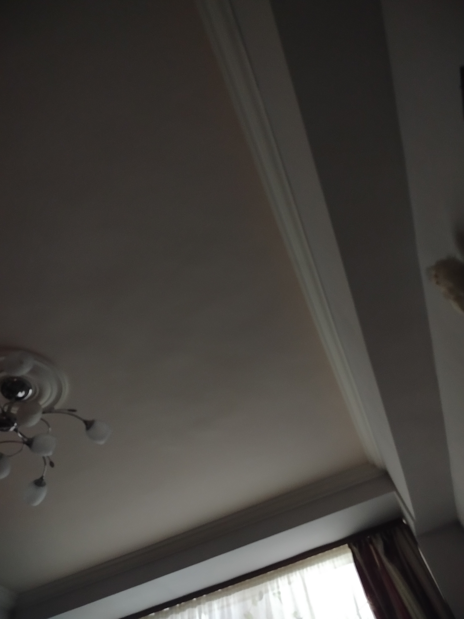 The image shows a view of an interior ceiling with a decorative chandelier in the bottom left corner and a window with curtains along the bottom edge. The ceiling is painted a light color. There is trim molding along the edges of the ceiling. A wall or beam is visible on the right side. The location is unknown, and there is no indication that this image was taken in Yerevan, Armenia.