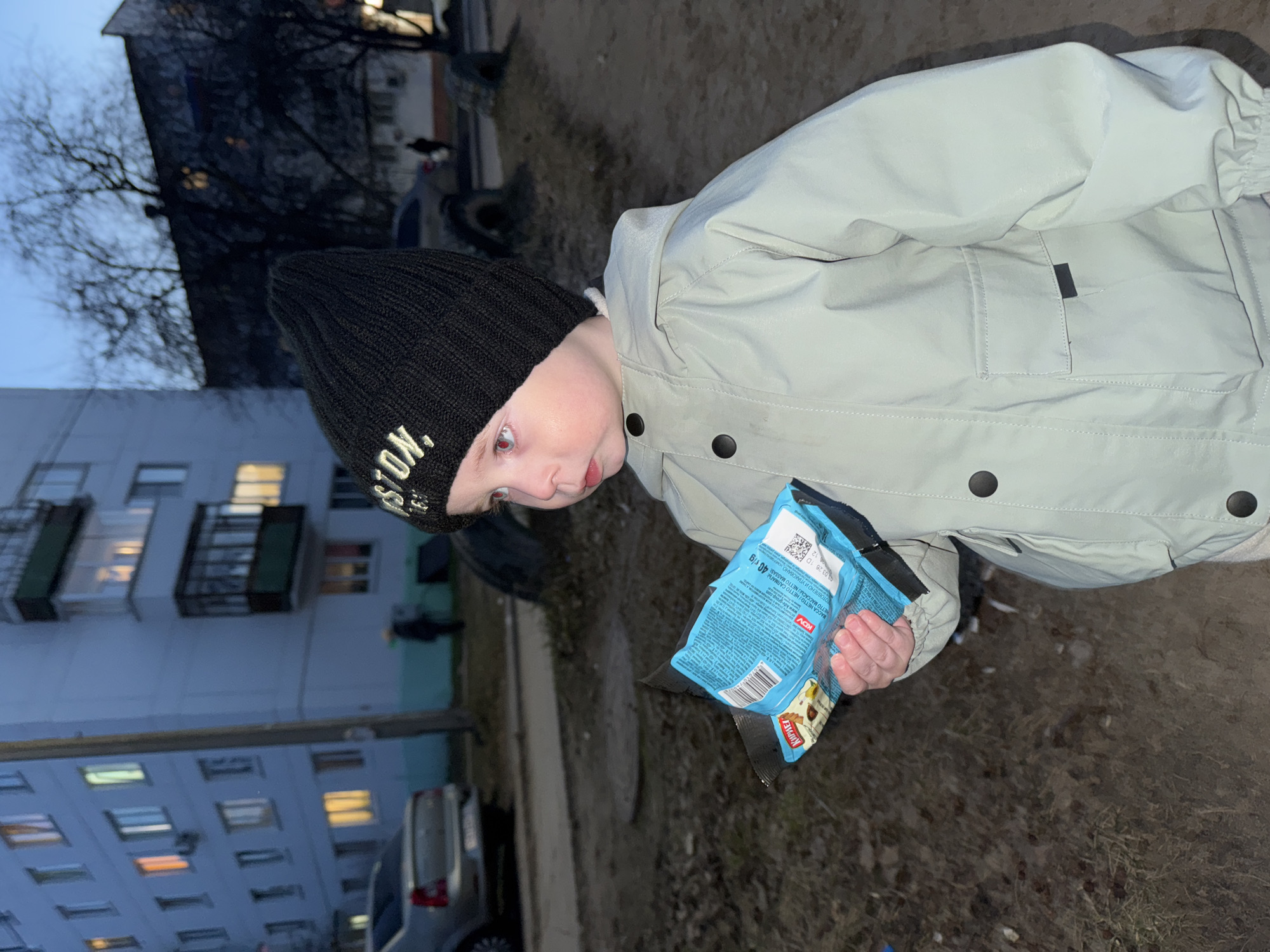 A young child wearing a black knit hat and a light-colored jacket stands outside holding a package of "Kupetz" wafer snacks. The child is positioned in front of a weathered, multi-story building, likely an apartment block, which is common in Chaikovsky, Russia. The lighting suggests it is late afternoon or early evening. In the background, there are parked vehicles and trees. The ground appears to be a mixture of dirt and grass.