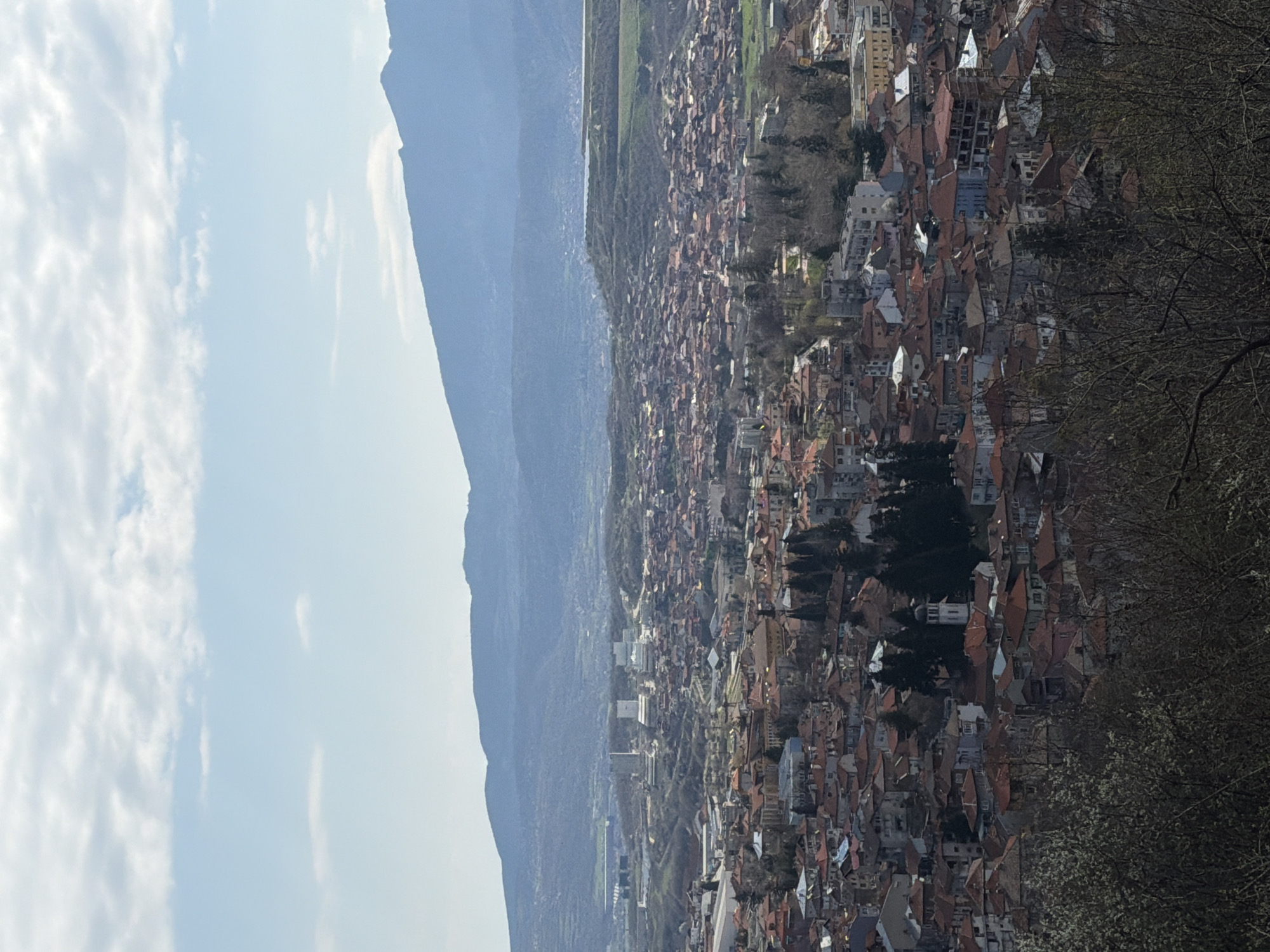 An aerial view depicts the city of Sveti Konstantin, Bulgaria, extending from the base of a mountain range toward the sky on the left. The sky contains scattered clouds. The cityscape consists of dense arrangements of buildings with terracotta roofs. Trees punctuate the urban landscape. In the foreground on the right is foliage.