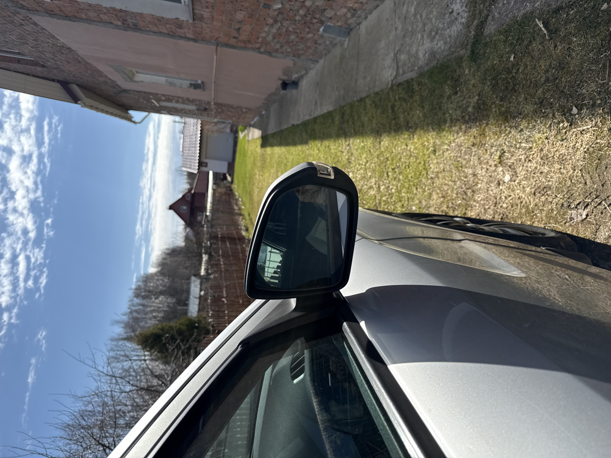 The image depicts a silver car parked on the side of a road in a residential area. The side mirror of the car is visible, reflecting the surrounding environment. A brick building is located on one side of the road, and a grassy area on the other. A narrow concrete path runs along the side of the building. In the distance, a red-roofed house is visible beyond bare trees under a partly cloudy sky. The location is specified as Адам, Russia.