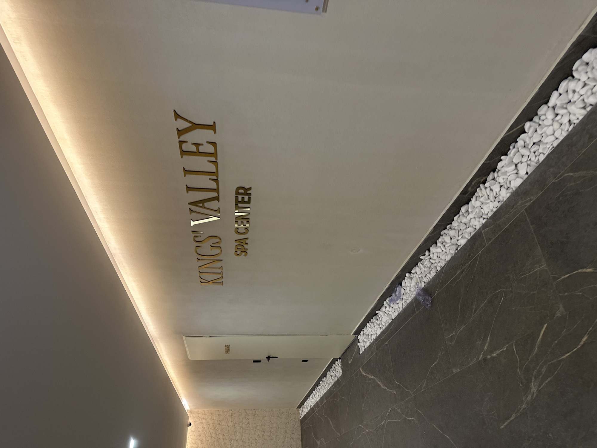 This is an interior photograph showcasing a modern and elegant spa entrance or hallway, located in Kazanlak, Bulgaria. The main focal point is a light-colored wall (appearing almost white or light beige) adorned with prominent, golden, three-dimensional letters. The larger text reads "KINGS' VALLEY", with "SPA CENTER" displayed below it in a smaller font.
Above and running parallel to this text, a strip of recessed lighting casts a warm, soft glow downwards, illuminating the wall and accentuating the golden letters. To the right side of the image, a dark, veined marble surface, possibly a floor or a lower wall, features a decorative channel filled with numerous smooth, white pebbles. This design element adds to the serene and natural aesthetic common in spa environments.
Below the main text and lighting, a plain white door is visible. Attached to this door is a small, rectangular sign that clearly reads "PRIVATE". The overall aesthetic is clean, sophisticated, and tranquil, characterized by the interplay of light and dark tones, natural textures, and subtle illumination. There are no people visible in the image, emphasizing the quiet and inviting atmosphere of the facility. The lighting indicates an indoor setting, and the decor suggests a high-end wellness establishment.
