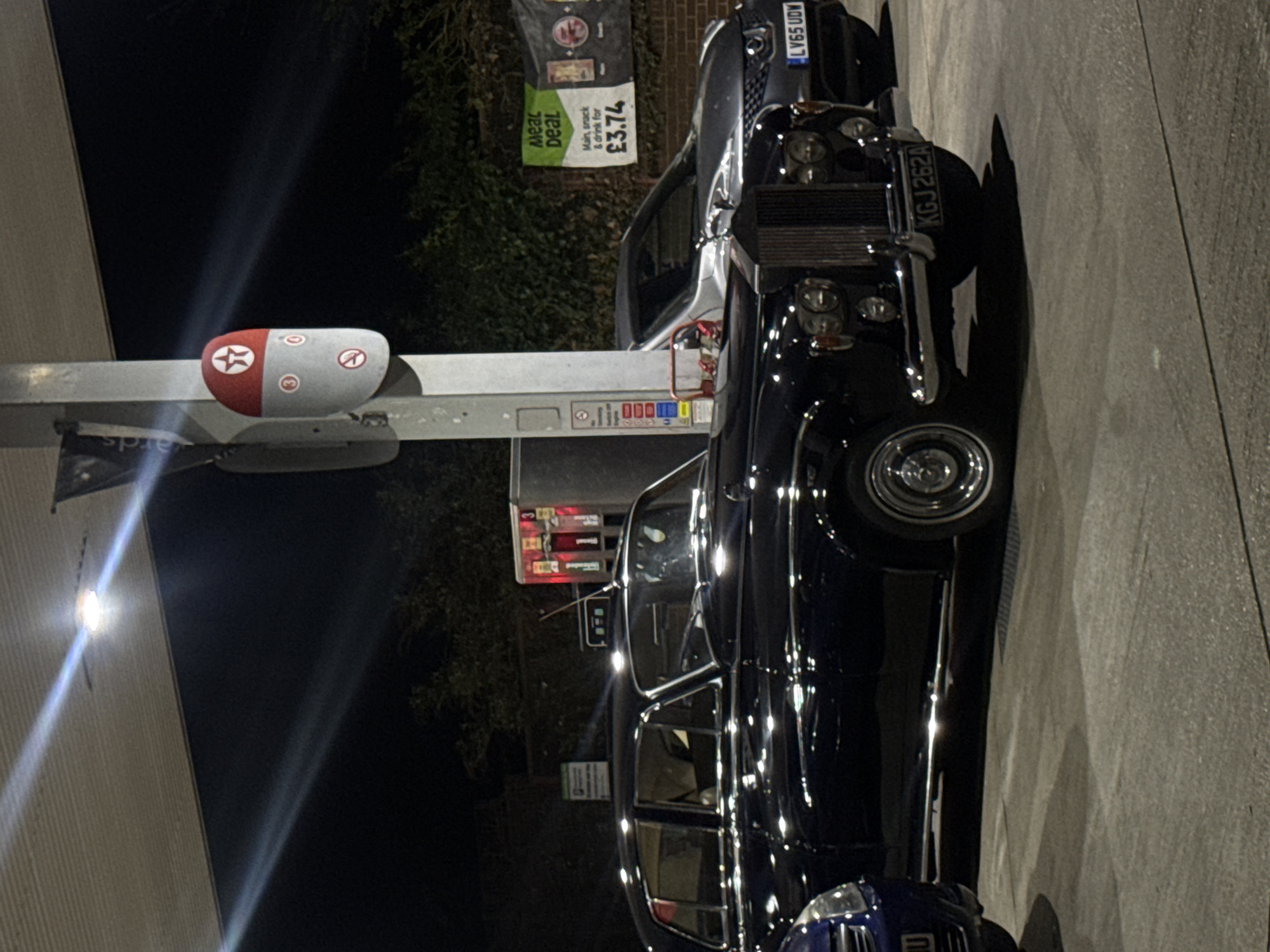 The image shows two luxury vehicles parked at a gas station, specifically a Texaco, at night. A Rolls Royce, with the license plate KGJ 262A, is prominently featured. Adjacent to it, there is another high-end vehicle, possibly another Rolls Royce or Bentley, with license plate LV65 UDW. A sign advertising a meal deal is visible, priced at £3.74. The gas station's pumps are visible. The location is likely somewhere in the UK due to the British number plates and currency, and is assumed to be the city of London, United Kingdom.