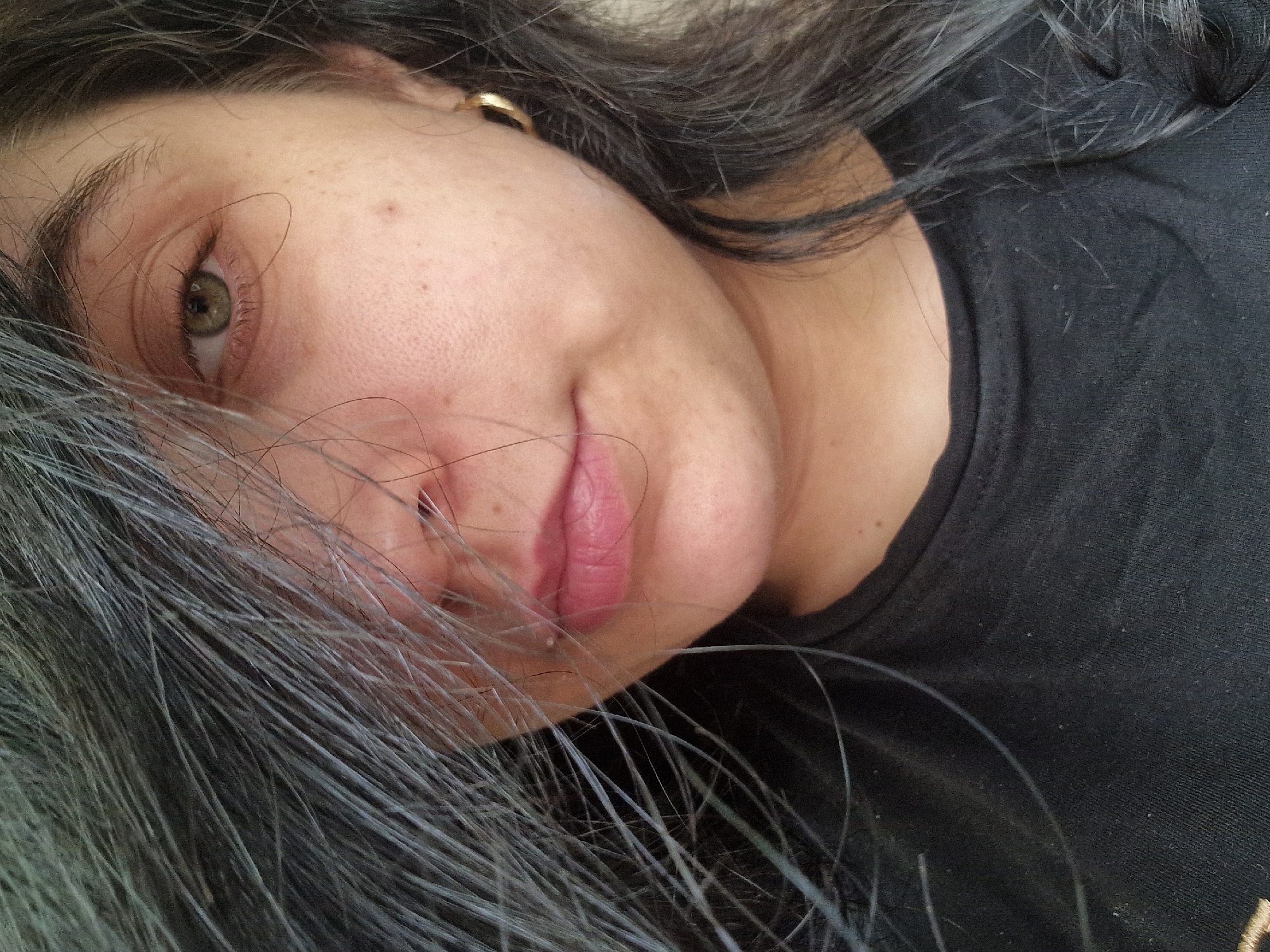 A close-up, oblique portrait of a fair-skinned individual with dark hair, positioned at an angle with the head slightly tilted. The person's face is partially obscured by strands of hair. They have an eye visible, and their lips are colored in a pink hue. The individual is wearing a black top. An earring is visible in their ear. The location is not identifiable from the image.