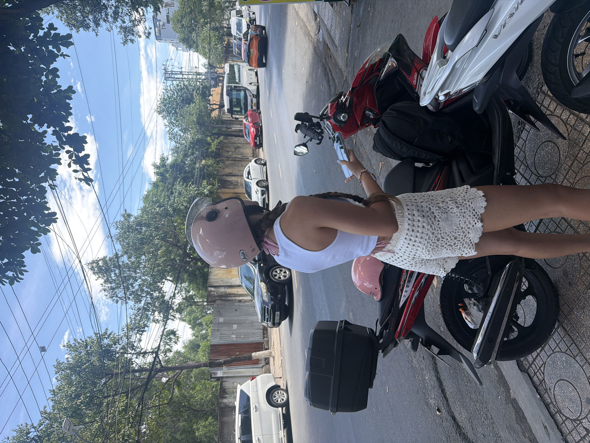 A young woman, facing away from the camera, stands beside a red scooter parked on a sidewalk in Nha Trang, Vietnam. She wears a white crop top, a pink scarf draped around her neck, and white crochet shorts. Her hair is styled in braids that hang down her back. On her head is a pink helmet. She is holding a smartphone in her right hand. Another pink helmet rests on the seat of the scooter. A black storage box is mounted on the rear of the scooter. The scooter is parked next to a white scooter. In the background, a black SUV is visible, along with other vehicles, trees, utility poles with wires, and buildings. The sky is blue with scattered clouds. The street is paved, and the sidewalk is tiled.