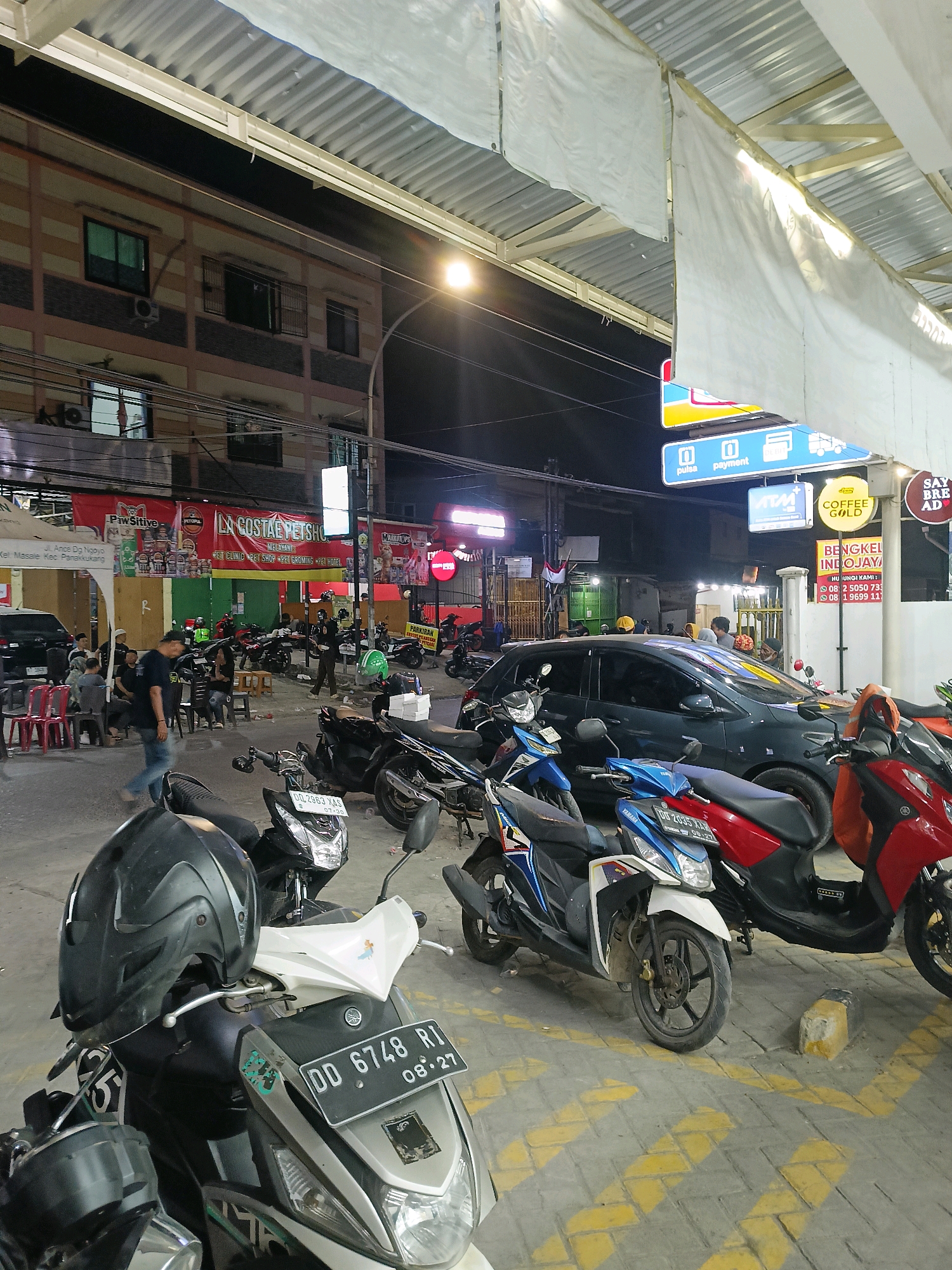 This is a medium shot, eye-level, horizontal photograph taken at night of a street scene in Makassar, Indonesia. The foreground is dominated by several parked motorcycles, with a black helmet resting on the handlebars of a white and gray scooter. The license plate of this scooter reads "DD 6748 R1 08.27". To the right, a red scooter is parked. In the midground, a dark gray sedan is parked, surrounded by more motorcycles. People are visible on the left side, some seated on chairs, others standing and walking. A large red banner advertising "LA COStAE PET SHOP" hangs prominently across a building in the background. The building itself appears to be a multi-story structure with balconies and windows. A street light illuminates the scene from the upper right. Signs for "pulsa" and "payment," "ATM," and "COFFEE GOLD" are visible above the businesses. The ground is paved with interlocking bricks marked with yellow directional arrows. The overall impression is of a busy commercial street at night, likely a food or service area.