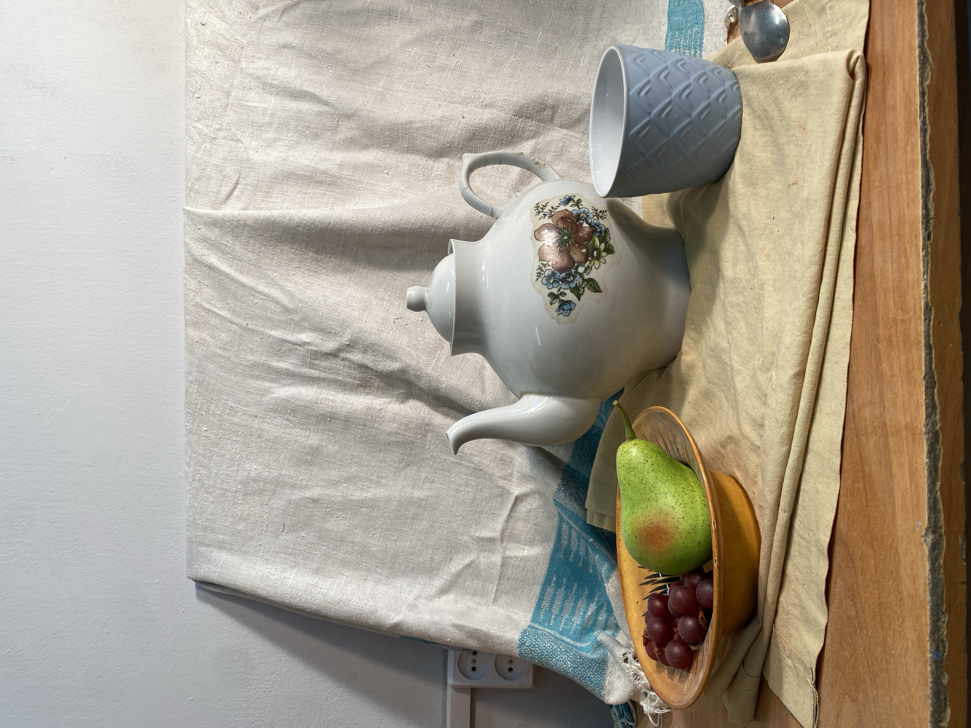 The image shows a still life arrangement on a textured surface, likely a draped fabric, with a portion of a wall and a wooden surface visible. The items include a white teapot with floral decoration, a light blue cup with a textured pattern, a wooden bowl containing a green pear and red grapes, and a silver spoon. A blue and white patterned cloth is also present. The scene is lit with soft, natural light, casting shadows on the left side. An electrical outlet is visible on the lower left side of the image. There is no evidence to indicate this image was taken in Yekaterinburg, Russia.