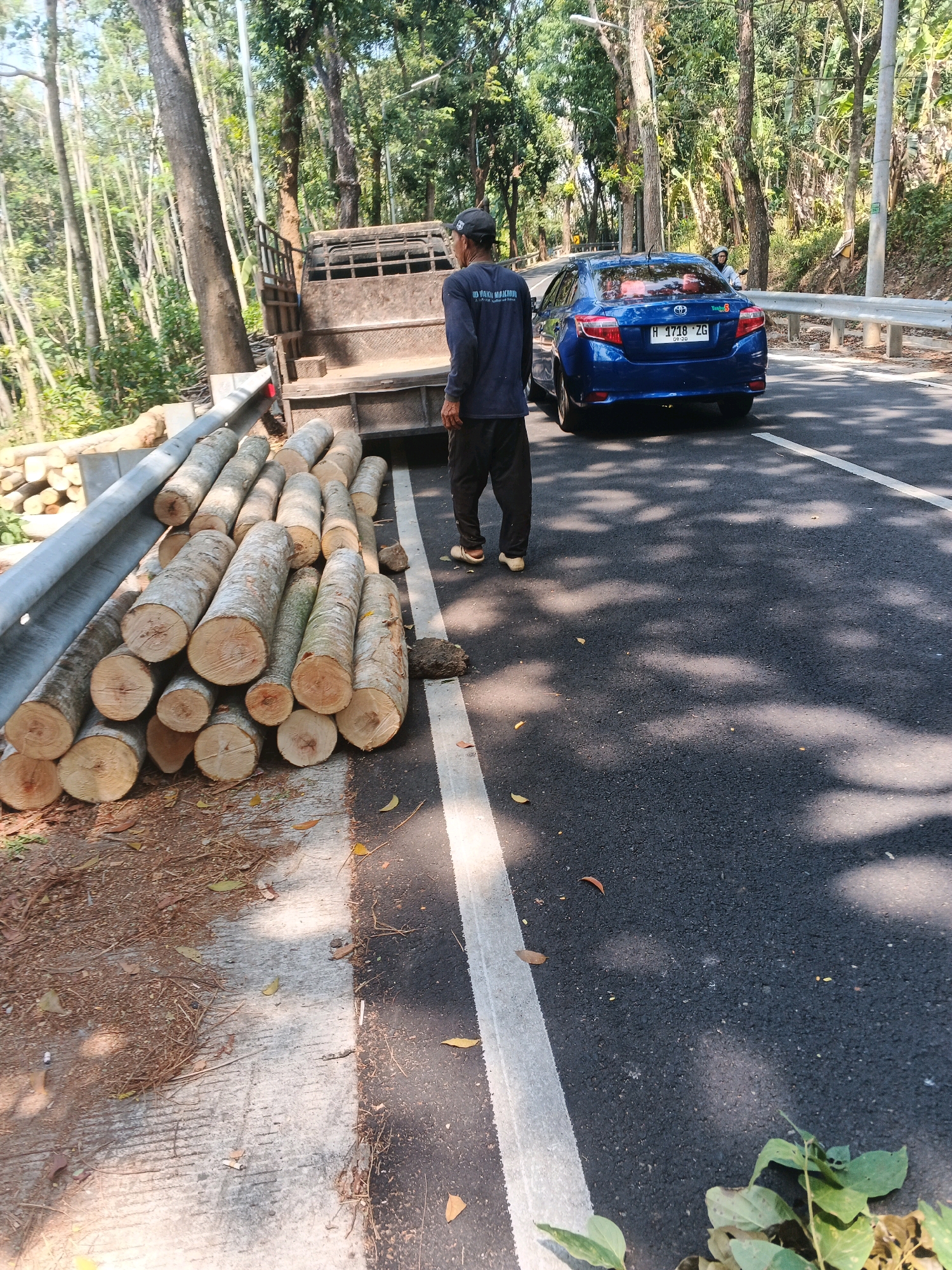 In Bandarjo, Indonesia, a man in a dark blue shirt and black pants stands next to a pile of cut logs on the side of a paved road. The logs are stacked on a concrete surface adjacent to a metal guardrail. Behind the logs, a weathered truck is parked. Further down the road, a blue sedan is driving, its license plate reading "H 1718 ZG". The area is lined with trees, suggesting a rural or semi-rural setting. The lighting indicates it is daytime, with shadows cast on the road.