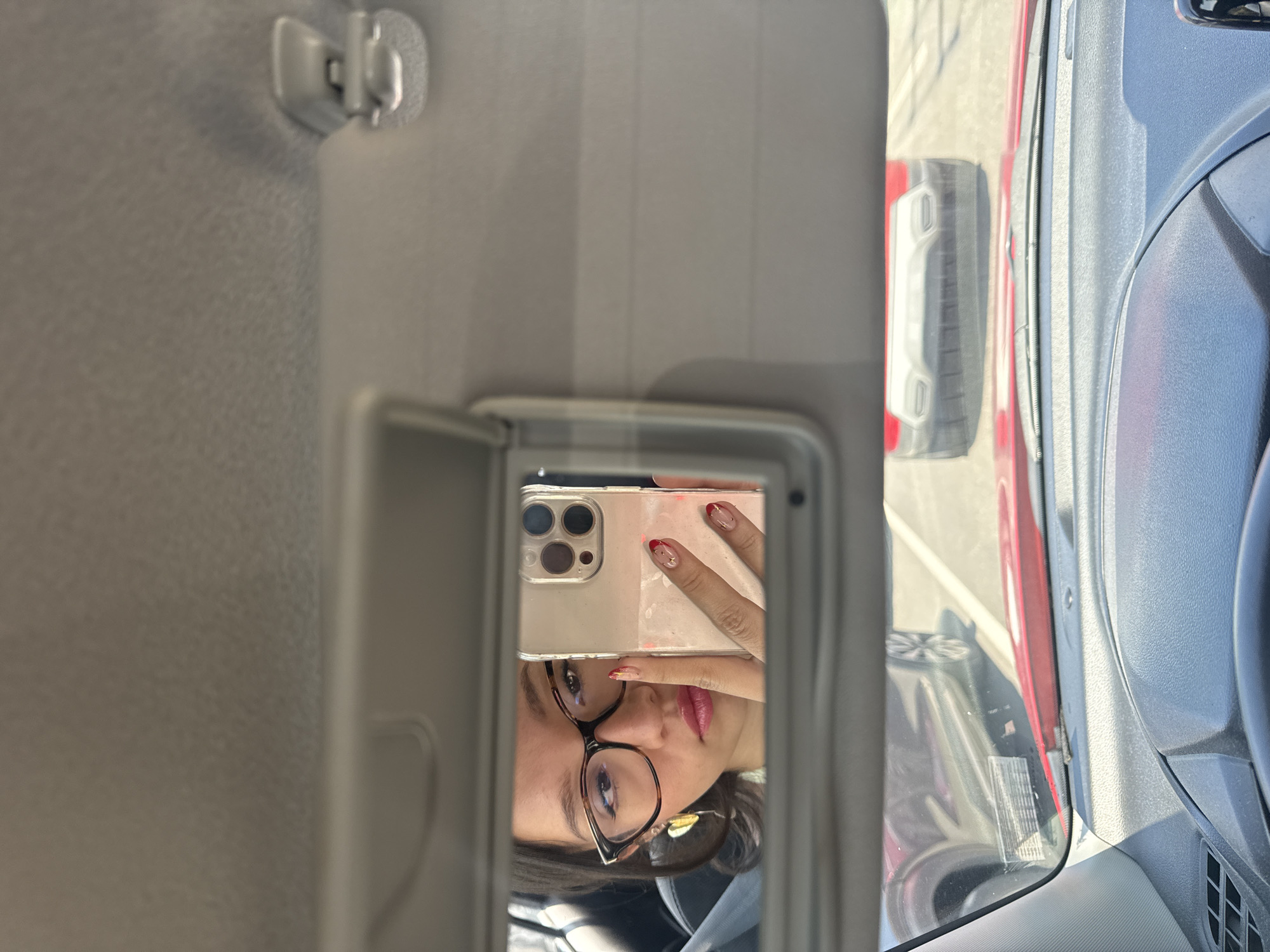 An inverted, eye-level close-up shows a person taking a selfie reflection in a car's open vanity mirror. The person's face, partially obscured by a smartphone, is centered in the mirror, looking directly into the camera. They wear tortoiseshell-framed eyeglasses and pink lipstick, with dark hair visible around their face. Their right hand holds a light-colored smartphone with a clear case and multiple camera lenses, positioned to capture the reflection. The person's nails are manicured with red tips and a subtle gold design.
The mirror is set within a light-colored car sun visor, which is open and angled downwards. Above the mirror, the car's light grey headliner and the attachment point for the sun visor are visible. To the right of the mirror, the dark grey dashboard and part of a steering wheel or door panel are visible through the car window.
Outside the car, under bright daylight, parts of a red vehicle are visible, including a prominent grey or silver bumper/trim and a tire, indicating another vehicle is parked nearby. The bright lighting suggests it is daytime and likely sunny. No discernible text is visible in the image. The image is taken inside a vehicle, likely in San Andrés Cholula, Mexico.