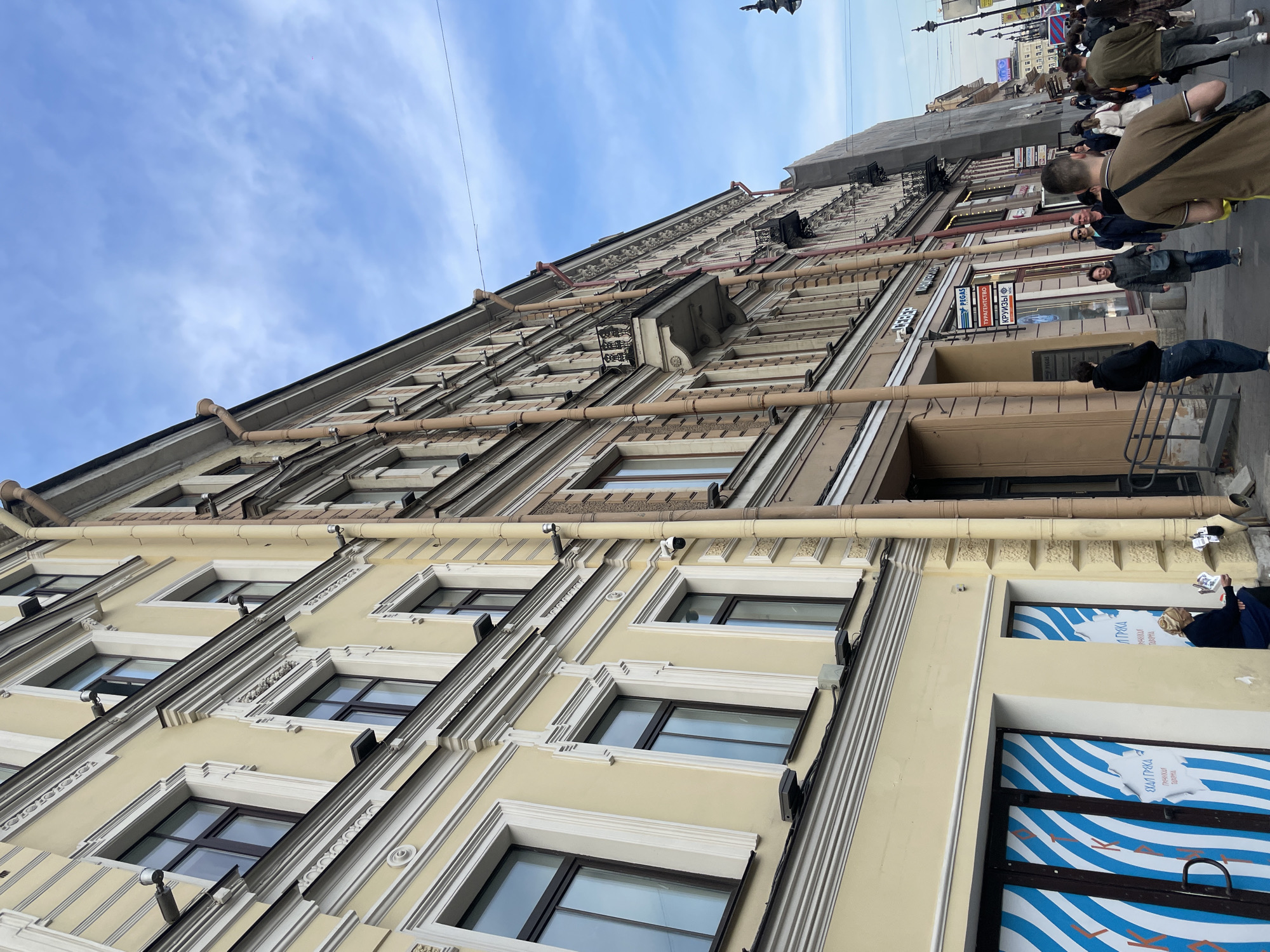 The image depicts a street scene in Saint Petersburg, Russia, focusing on a multi-story building with architectural details such as decorative window frames, prominent drain pipes, and ornate moldings. The building has a yellow facade. Street-level storefronts feature blue and white striped designs and signage in Russian. Pedestrians are walking along the sidewalk in front of the building. The sky is partly cloudy.