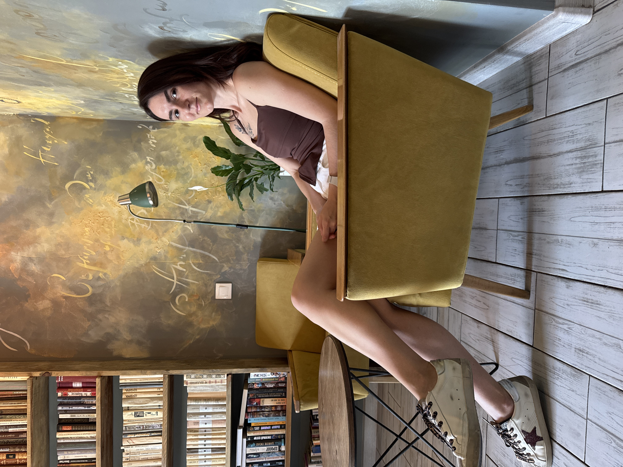 Here's a detailed description of the image:
**Scene:** Indoor setting in what appears to be a bookstore or library cafe.
**Location:** Likely Tyumen, Russia, based on the Cyrillic writing on the books on the shelves.
**People:** One woman is present. She is sitting in a mustard-yellow armchair. She is wearing a dark brown tank top, white shorts, and white sneakers with a star design. She has dark hair and is looking directly at the camera.
**Objects:**
* The woman is sitting in an armchair.
* Behind the armchair, there's a tall bookcase filled with books. Many of the books have titles in Cyrillic script.
* A small, round table is positioned in front of the armchair.
* There's a floor lamp with a green shade illuminating the area.
* A houseplant is placed next to the armchair.
* The walls have a textured paint effect with writing (possibly artistic calligraphy) in a golden color.
* The floor appears to be made of light-colored wooden planks.
**Actions/Interactions:**
* The woman is posing for the camera.
**Environment:** The overall atmosphere seems cozy and inviting. The presence of books suggests a place where people can relax and read. The lighting is soft, contributing to a calm mood. The interior design is somewhat eclectic.