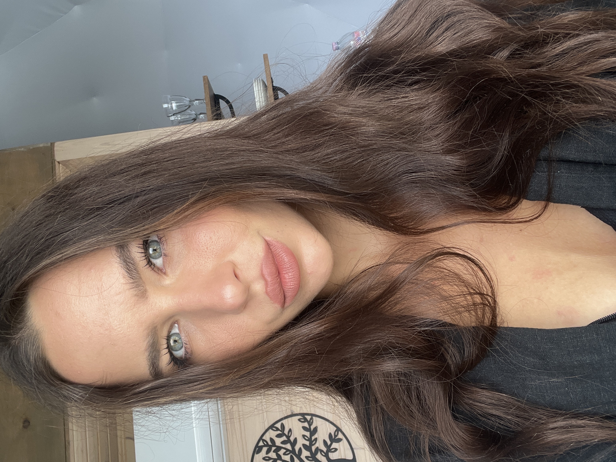 A young woman with long, brown, wavy hair and light green eyes is lying down facing the camera. She is wearing a black top. The background includes a wooden surface with a decorative tree cutout, wooden shelves holding wine glasses, and a light-colored wall. The location is not specified in the image.