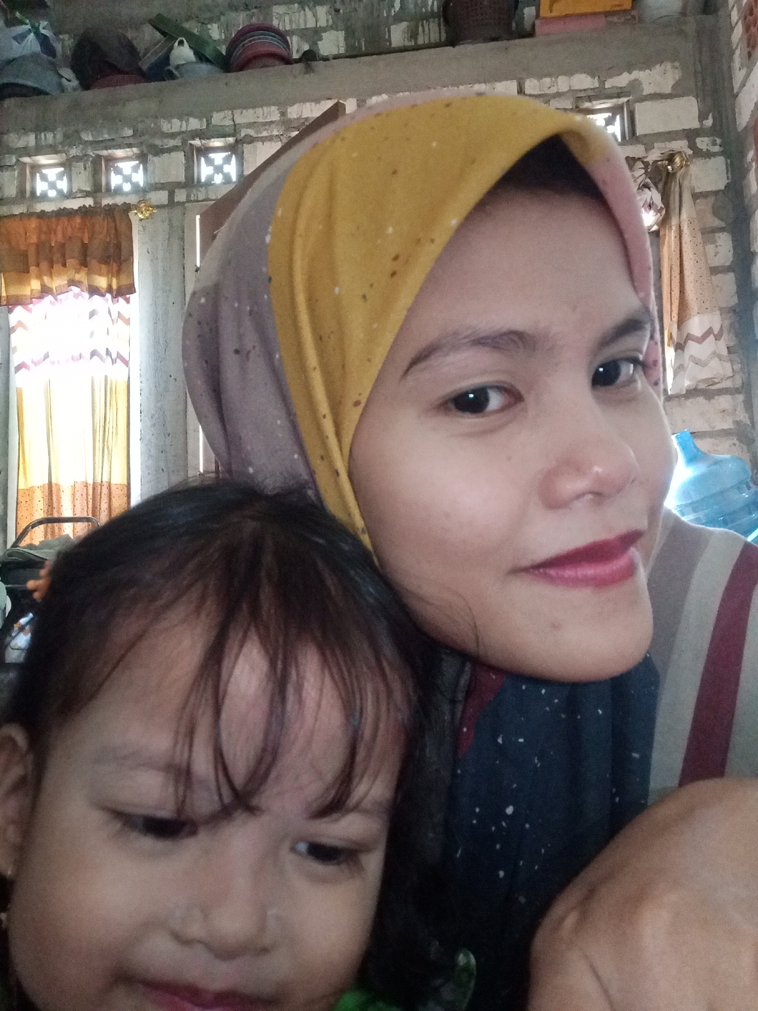 The image is a selfie taken by two individuals, an adult female and a young child, indoors. The female, on the right, is wearing a hijab that transitions from yellow to pink and beige, and a striped shirt. She is looking directly at the camera, with makeup on her lips. The child, on the left, is out of focus and appears to be looking down. The background shows a wall constructed of hollow concrete blocks, with ventilation bricks. There are also curtains visible, and a large blue water dispenser in the background. The overall impression is of a casual, domestic setting. The context of Juwana, Indonesia, is not discernible from the visual elements within the image itself.
