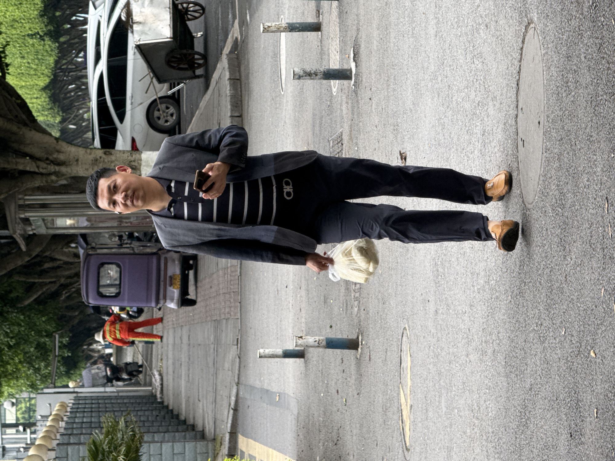 A man in a dark suit, striped shirt, and brown shoes walks on a paved road in Jiangjin, China. He holds a smartphone in his left hand and a plastic bag in his right. The road is lined with bollards and a manhole cover. A worker in an orange jumpsuit sweeps the sidewalk on the left, next to a small purple vehicle. In the background are trees, a white car with a food cart attached, and stairs.