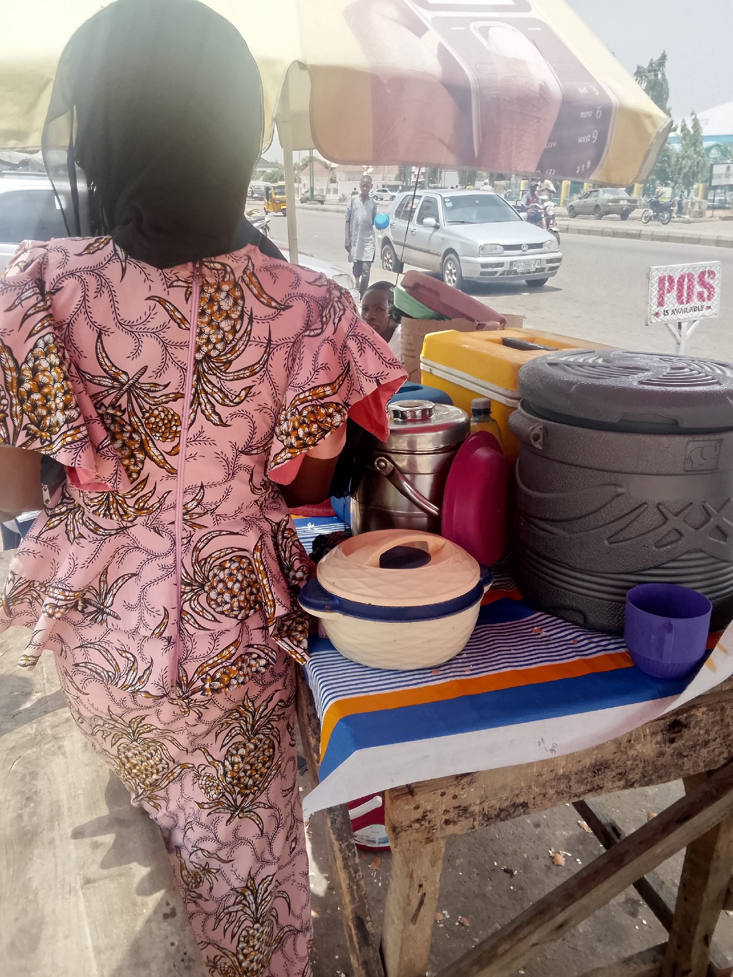 An outdoor scene, likely Pantami, Nigeria. A woman with a black hijab and a pink dress with pineapple patterns stands beside a wooden table covered with a blue and white striped cloth. On the table are containers: a blue and beige bowl with a lid, a stainless steel thermos, a pink plastic bottle, a yellow cooler, a large gray cooler, and a purple cup. A sign reading "POS IS AVAILABLE" is visible. A light-colored umbrella with a phone keypad design shades the area. In the background is a street with a silver car and pedestrians. A child's face is partially visible near the table.