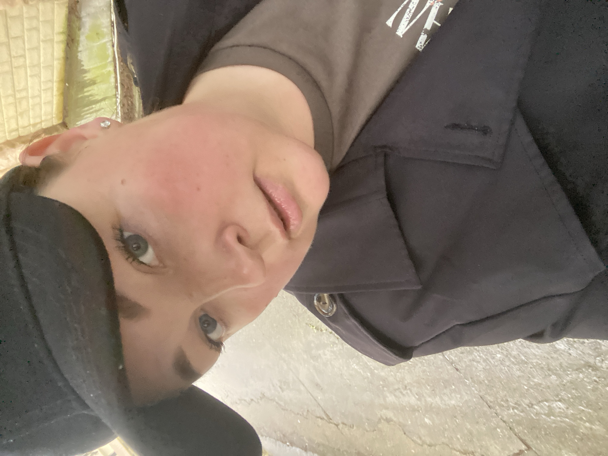A headshot-style image depicts a person, likely female, located in Sochi, Russia. The individual is viewed from a slightly elevated angle, looking directly into the camera. She wears a dark, possibly black, baseball cap and a dark, presumably navy or black, collared jacket or trench coat with a visible button on the right shoulder epaulet (viewer's left). Underneath, a brown or olive-drab t-shirt displays a partial white graphic or text on its front. The subject exhibits a fair complexion with noticeable redness on the cheeks, light blue-grey eyes, dark eyebrows, and a small, round, silver-colored stud earring in her right earlobe (viewer's left). Her lips are naturally colored and slightly parted. The immediate background shows a light-colored, textured ground surface, possibly concrete or pavement, appearing damp or reflective. Further back and to the subject's right (viewer's left), a light-colored brick or block wall is visible, exhibiting signs of moisture and green biological growth along its lower section.