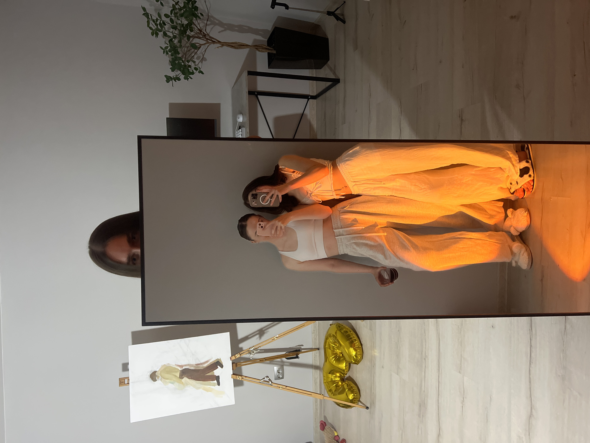 In a domestic setting, two individuals are captured in a reflected image within a large, full-length mirror. The mirror occupies the foreground and a significant portion of the frame, obscuring the viewer's perspective of the room's entirety. The reflection shows two young women, both dressed casually in what appears to be loungewear: one in a white crop top and light-colored sweatpants, holding a cup in her left hand, and the other in a similar white top and flowing trousers. Both are holding smartphones up, presumably taking selfies, and their faces are partially visible as they focus on their devices.
Behind the mirror, a section of the room is visible. To the left, an easel stands with a painting on it, depicting two figures in muted tones. Nearby, large, gold-colored foil balloons are arranged on the floor. The background includes a wall, a desk with a computer monitor, a mouse, and a potted plant in a dark planter. A tripod is also visible to the right. The floor appears to be light-colored wood laminate. The overall lighting suggests an indoor environment, possibly an apartment, with a warm, orange-toned light source casting prominent shadows and highlights, particularly on the lower half of the reflected figures and the floor. There is no specific indicator within the image to pinpoint the location as Ternopil, Ukraine.