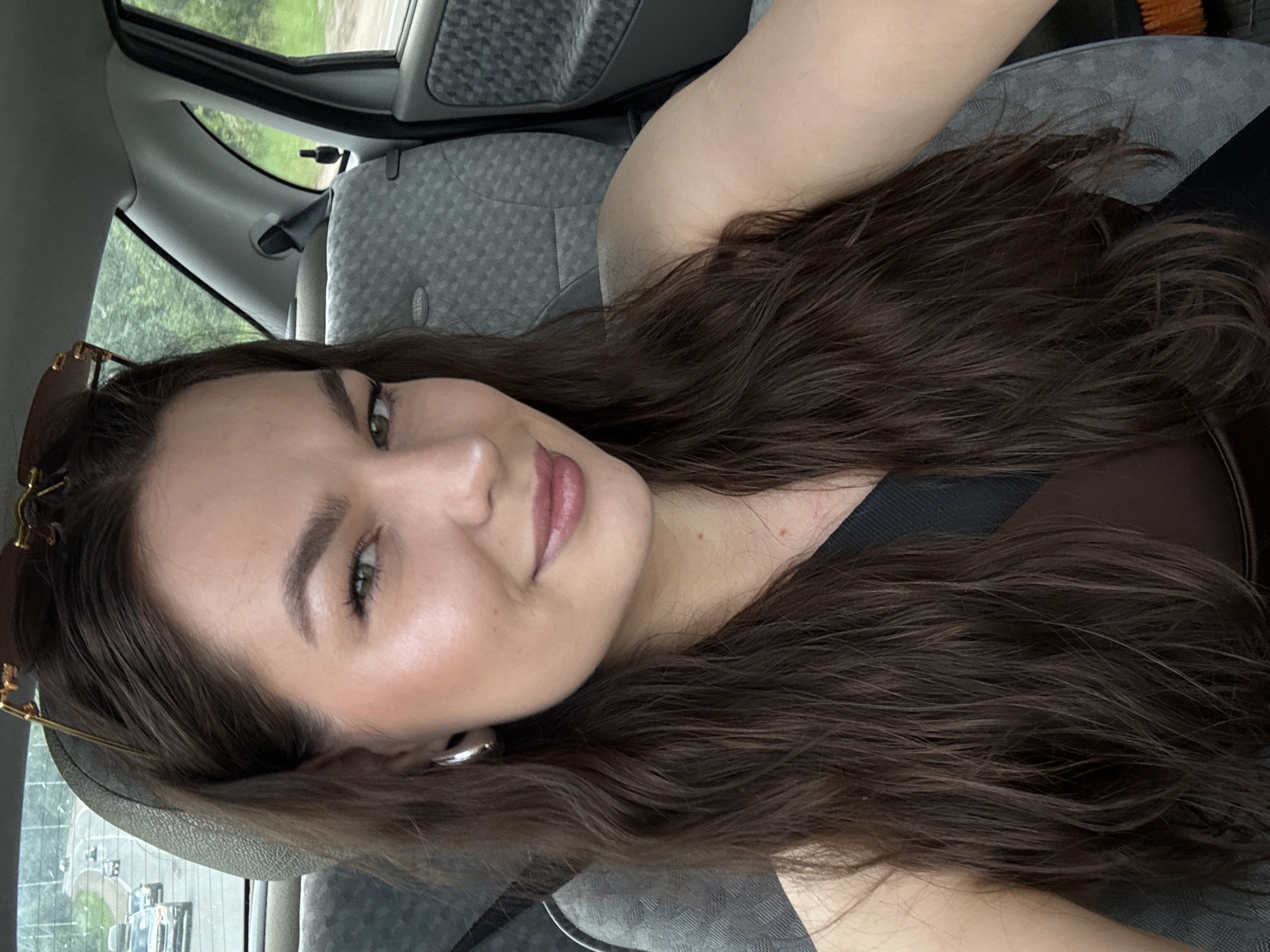 A young woman with long, dark brown wavy hair is taking a selfie in the passenger seat of a car. She is wearing a dark brown top and large silver hoop earrings. Sunglasses are perched on top of her head. Her makeup is visible, including winged eyeliner and lip gloss. The car's interior is visible behind her, including a grey patterned seat and the headrest. Outside the car window, a road with several vehicles is visible, along with trees and greenery, suggesting a rural or suburban setting. The overall scene appears to be taken during daylight. The location is identified as Chaikovsky, Russia.