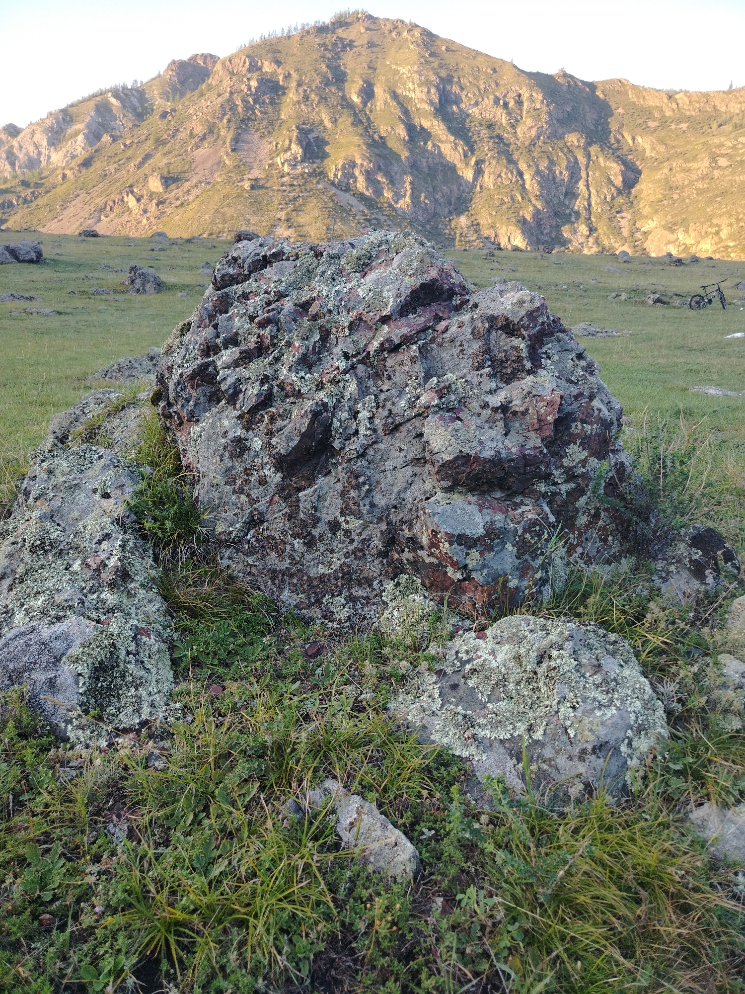 The image shows a large, weathered boulder covered in moss and lichen in the foreground, surrounded by green grass and smaller rocks. In the background, a mountain range with sparse vegetation rises against a clear sky. The lighting suggests it is either early morning or late afternoon, casting long shadows. A bicycle is visible in the middle ground on the right side of the image. The scene appears to be a natural landscape, likely in a rural or mountainous area, and is not specifically located in the city of Iodro, Russia, as no identifiable landmarks or urban features are present.
