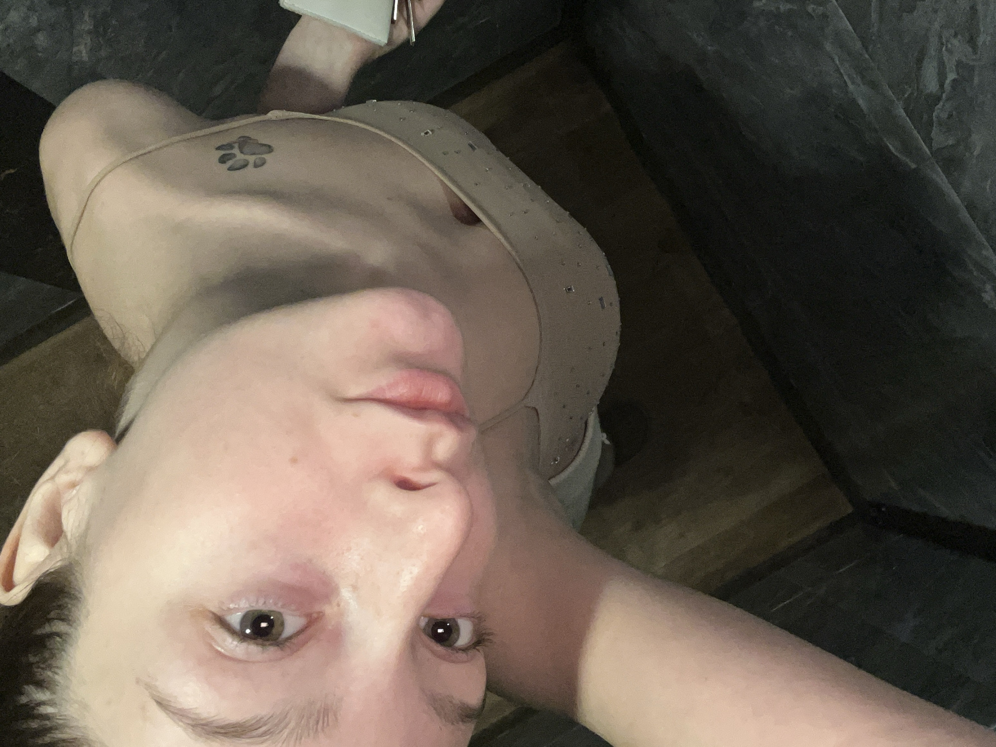 A high-angle selfie captures a light-skinned individual, likely an adult female, looking directly into the camera. Her face, including her eyes, nose, and slightly parted lips, is prominent in the foreground, angled upwards. Her dark brown eyes are open and focused, and her expression is neutral. A faint redness is visible on her cheeks, and her left ear is clearly visible.
She is wearing a light-colored, possibly beige or nude, top or dress with thin straps. The fabric of the top appears to have small, possibly reflective, embellishments or details. A dark paw print tattoo is visible on her upper left shoulder/collarbone area.
The background consists of dark, textured surfaces, suggesting an indoor setting. On the right and upper right, dark grey or black surfaces, resembling stone or rough concrete, form a wall or corner. The lower left and bottom parts of the background reveal a lighter, dark brown surface, possibly wooden flooring or a wooden wall panel. The overall lighting appears somewhat dim, creating a subdued atmosphere. No visible text is present. The scene is situated in Varna, Bulgaria.
