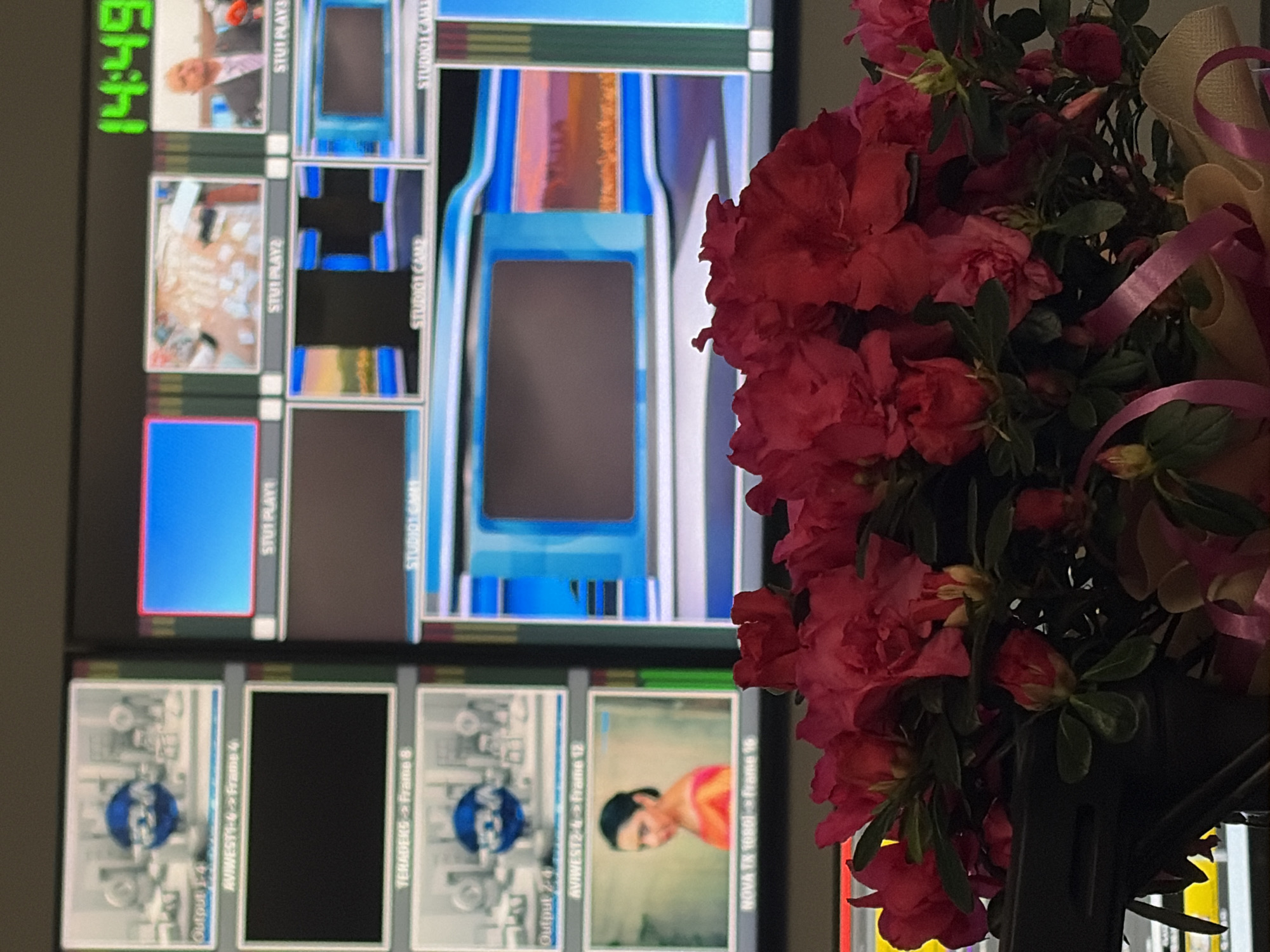 A vibrant bouquet of pink and red azalea flowers with lush green leaves, accented by a beige/gold and pink ribbon, occupies the right side of the frame, partially obscuring the background. The flowers appear to be in a dark pot or stand.
In the background, a large monitor displays multiple video feeds, indicating a broadcasting or media control room setting in Busmantsi, Bulgaria. The current time "14:49" is visible in green in the top left corner of the screen.
The monitor features various feeds:
- Top row includes a man in a suit (labeled "STUI PLAYE"), a bright, cluttered scene (labeled "STUI PLAY2"), a solid blue screen, and a blue and grey graphical display (labeled "STUDIOT CAM"). Other similar labels like "STUI PLAYS" and "STUDIOT CAME" are also partially visible.
- Bottom row shows technical graphical displays with text like "Output 1-4" and "AVIWEST1-4 Frame 4," a black screen labeled "TERADERS Frame 8," another technical graphic labeled "Output 2-4" and "AVIWEST2-4 Frame 12," and a feed of a woman with dark hair wearing an orange garment, labeled "NOVA TX 1080) Frame 16."
The overall scene combines the functional, technical environment of a broadcast monitor with a touch of natural beauty from the flowers, likely indicating a workspace.