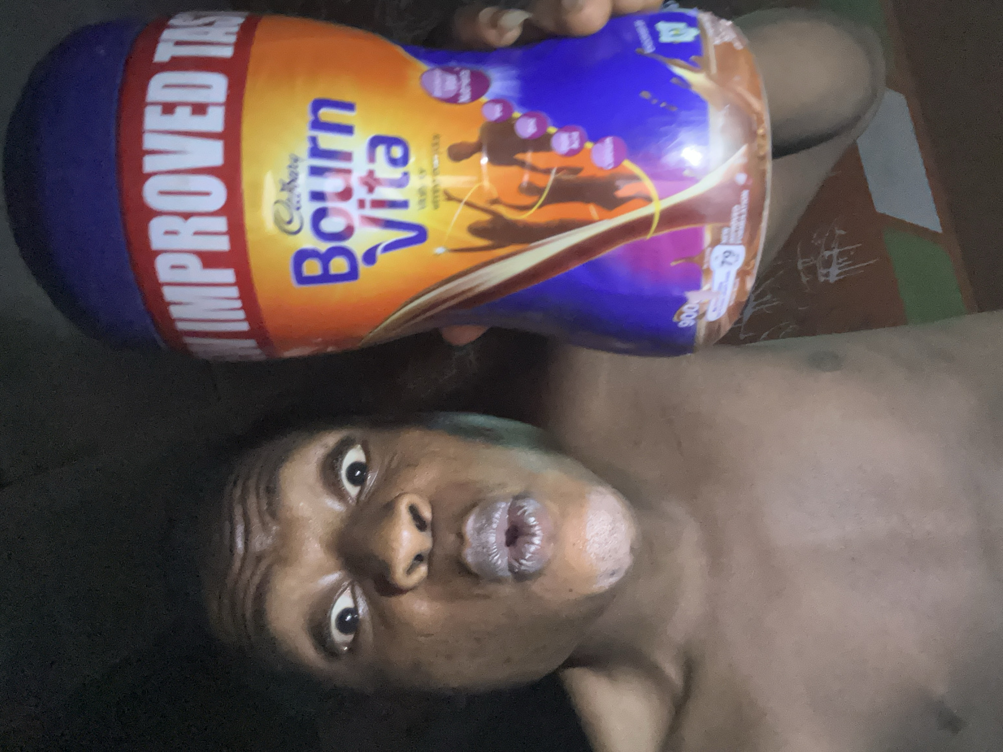 A person with dark skin lies on their back, holding a container of Cadbury Bournvita drink mix above their head. The container is predominantly orange and blue, with the words "Bournvita" prominently displayed. The person's face is visible, with wide eyes and pursed lips. The environment appears to be indoors. The location is unknown.