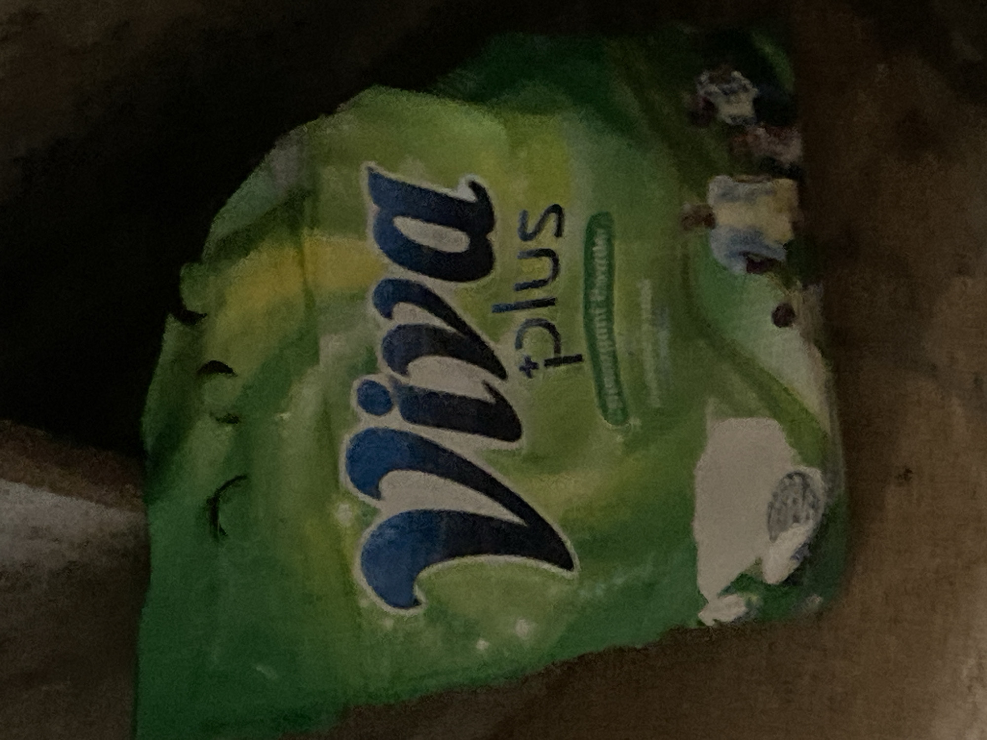 The image shows a green and yellow packet of "Viva Plus" detergent, with a faint illustration of people playing football. The packet is crumpled and appears to be a close-up shot, possibly taken indoors or in a shaded area. The lighting is dim, with some areas overexposed. The background is dark and out of focus. There are no specific landmarks or elements visible that would allow for pinpointing a location in Jauro Yinu, Nigeria. The image focuses solely on the product packaging.