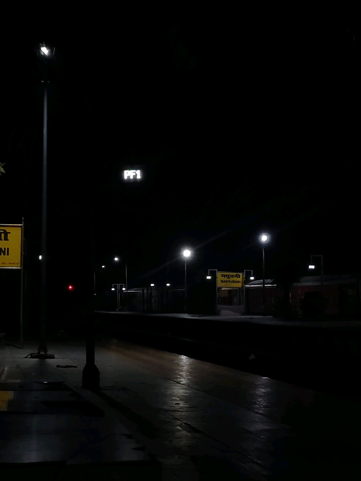 The image depicts a nighttime scene at a train station in Madhubani, India. A tall lamppost with a bright light stands to the left, casting long shadows. A yellow sign with text in Hindi and English, reading "MADHUBANI", is visible to the left of a platform. Another sign in the distance also displays "PF1". The platform is illuminated by several lampposts, with train tracks running alongside. A red signal light is visible on the platform. The ground appears to be wet, reflecting the light from the lamps. Buildings or structures are visible in the background, partially obscured by darkness. The overall atmosphere is quiet and still, characteristic of a deserted train station at night.