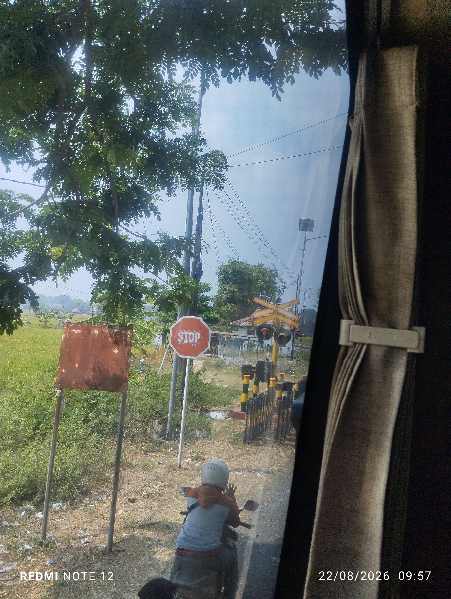 The image captures a street scene in Keras Wetan, Indonesia, as seen from inside a vehicle. A red octagonal stop sign is prominently displayed on the right side of the road. To the left of the stop sign, a rusted, square metal sign is mounted on two poles. A person wearing a white helmet is seated on a motorcycle, facing away from the camera. In the background, a railway crossing is visible with yellow and black striped barriers. The sky is bright and overcast, with power lines crisscrossing overhead. The area appears to be rural, with lush green fields on the left side of the road and some vegetation on the right. A curtain is visible on the right side of the frame, suggesting the image was taken from a window. The bottom of the image displays the text "REDMI NOTE 12" and the date and time "22/08/2026 09:57".