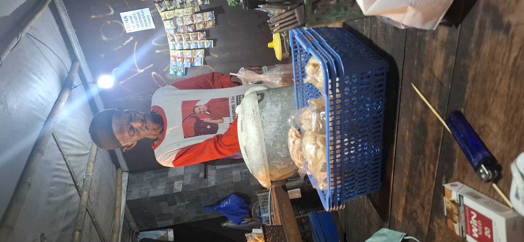 The image depicts an outdoor food stall in Colomadu, Indonesia. An elderly man wearing a dark skullcap and a red and white long-sleeved shirt with a picture of a politician and the text "Pramono, S.Sos" is standing behind a counter. In front of him, on a wooden table, is a blue plastic basket filled with bagged snacks. To the left of the basket is a pack of "Win Mild 20" cigarettes and a blue lighter. A wooden skewer runs through the lighter. In the background, shelves are stocked with various packaged food items and a sign lists food options. The stall appears to be illuminated by a bright light bulb. The overall setting suggests a humble, local business operating at night.