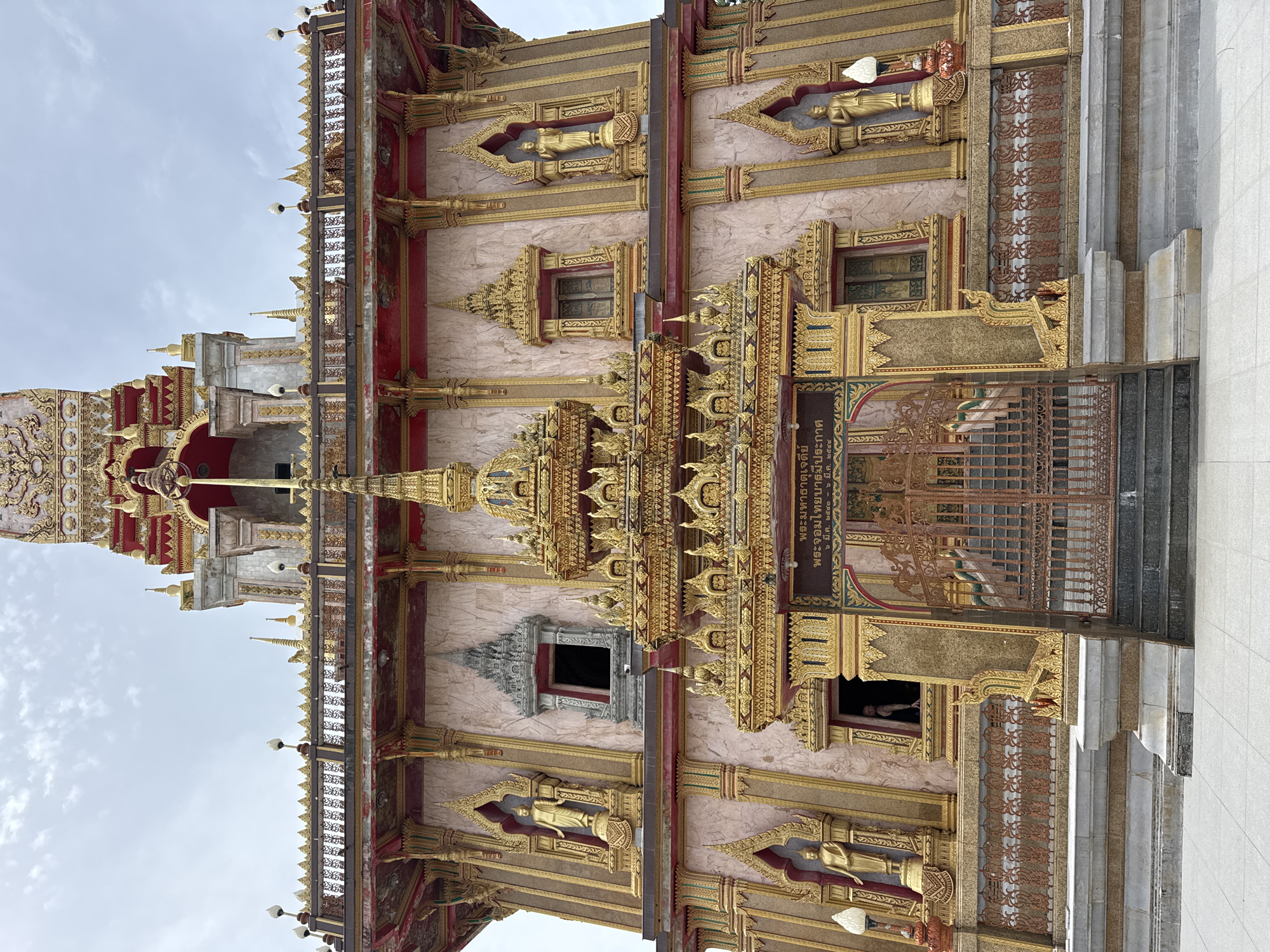 The image shows the exterior of the Chalong Temple, also known as Wat Chalong, located in Chalong, Phuket, Thailand. The temple is a Buddhist temple and is a prominent landmark. The architecture is ornate and features gold-colored decorations, statues of Buddha, and intricate carvings. The building has a light pink facade with gold trim. In the foreground, there is a wide tiled walkway leading up to a set of stairs and a large ornate metal gate. The gate is closed, with visible stairs behind it. Above the gate, there is a sign with Thai script. The sky is overcast. There are no people clearly visible in the image, though a silhouette of a person can be seen in a window to the left of the gate. The temple exhibits typical Southern Thai temple architectural style, with multi-tiered roofs and decorative elements.