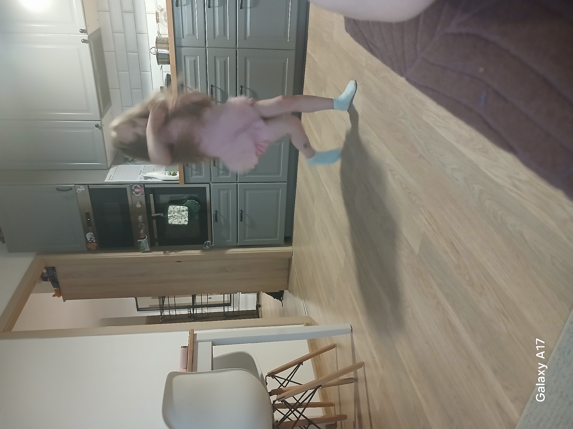 A blurry, low-light, indoor scene depicts a young girl in a pink dress and blue socks twirling in a modern kitchen. The kitchen features light gray cabinets and a wooden countertop. To the left, a white chair with wooden legs is partially visible, situated near a white table. In the background, a doorway reveals a glimpse of another room with a shelving unit. The floor is light-colored wood. The image is tagged "Galaxy A17" in the bottom left corner.