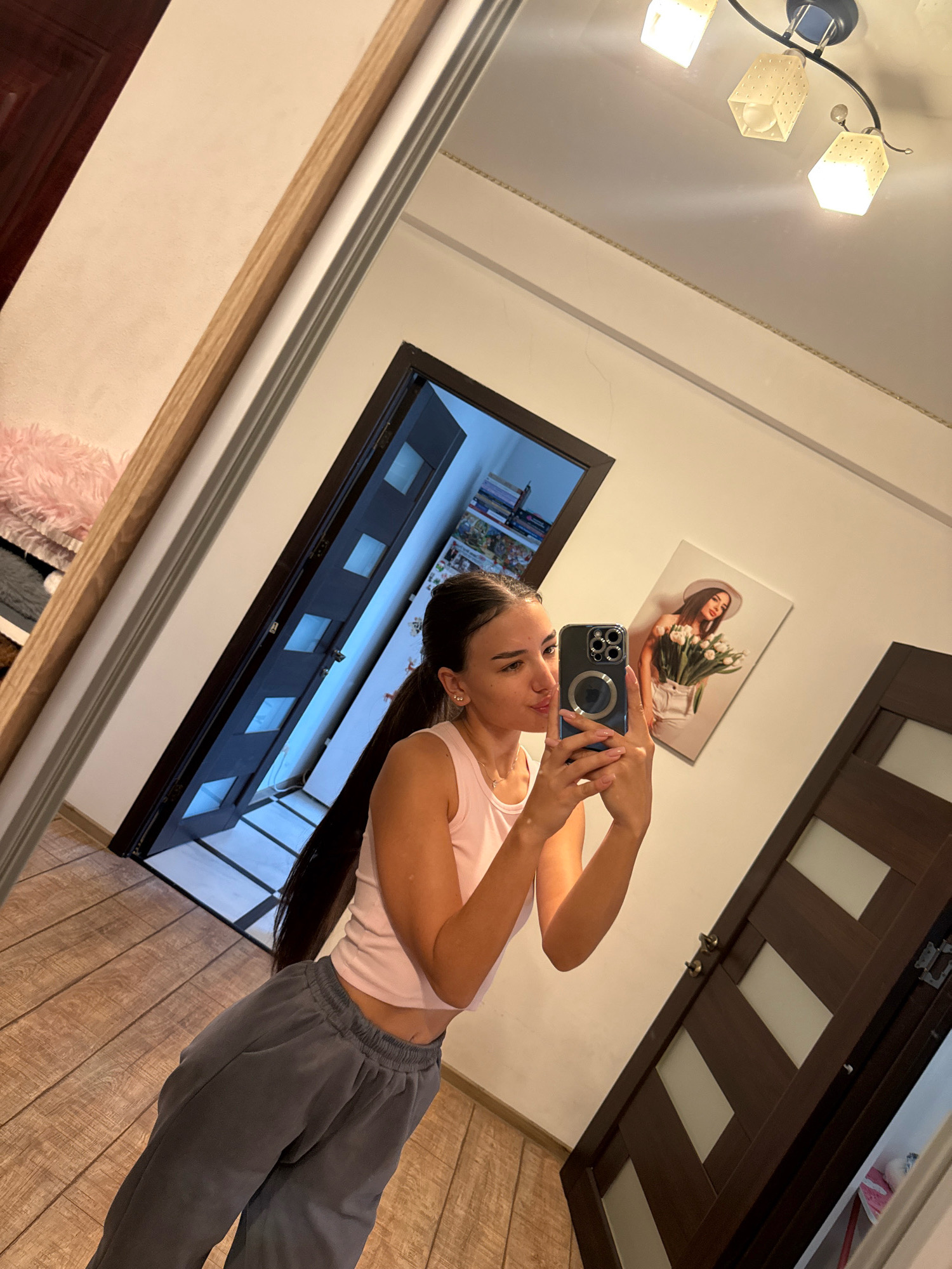 A young woman with long, dark hair tied into a ponytail is taking a mirror selfie in an indoor hallway. She is wearing a light pink tank top and gray sweatpants. The mirror reflects an open doorway with a dark-colored door, revealing a bright room beyond. On the wall opposite the mirror, a framed picture of a woman holding a bouquet of tulips is visible. A ceiling light fixture with three square lamps is also illuminated. The floor is covered with light brown wooden planks. The overall environment appears to be a residential interior, possibly an apartment or house hallway, but its specific location within Chișinău, Moldova cannot be determined from the visual information alone.