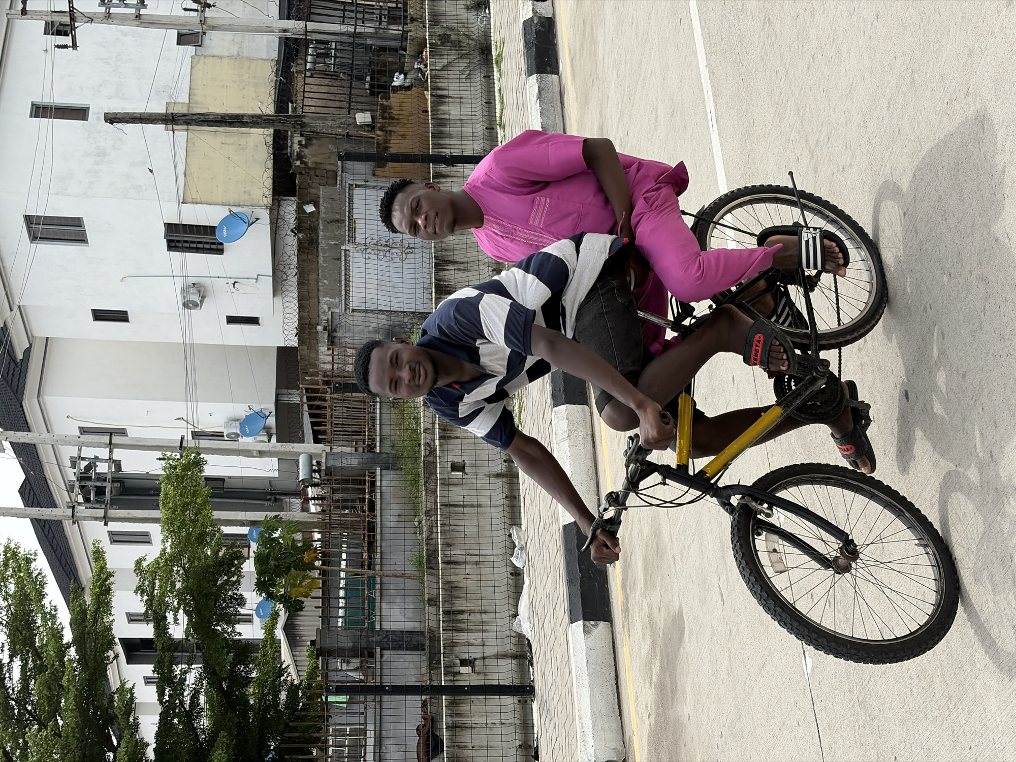 Two young men are riding a bicycle on a paved street in Igboefon, Nigeria. One man, wearing a striped shirt and dark shorts, is pedaling the bicycle. The other man, wearing a pink outfit, is riding on the back. Buildings and trees are visible in the background. A black and white curb runs along the edge of the street. A chain-link fence is visible on the left.