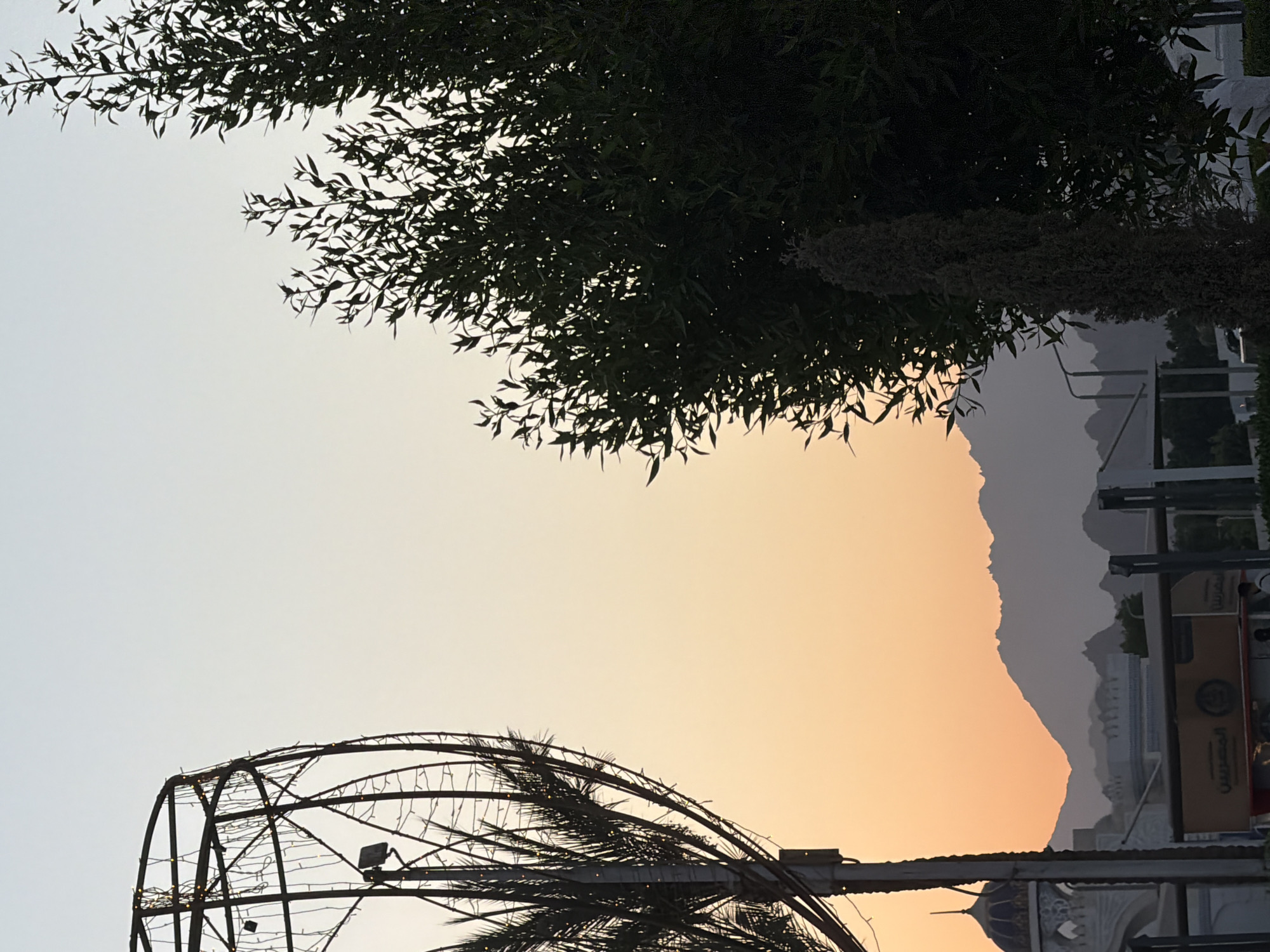 This media file shows an outdoor scene at dusk in Sharm El Sheikh, Egypt. Silhouettes of trees, including palm fronds, are prominent against a soft gradient sky transitioning from pale blue to warm orange hues, suggesting sunset. In the foreground, a large, dark, skeletal structure, possibly a decorative arch or frame adorned with small lights, is visible. A streetlight is attached to a pole supporting part of this structure.
In the background, hazy mountains form a silhouette against the colorful sky. Below the mountains, architectural elements of buildings are faintly discernible, including what appears to be a minaret on a dome and some signage. The overall impression is one of a calm, transitional moment between day and night in a scenic location. The lighting indicates it is either dawn or dusk.