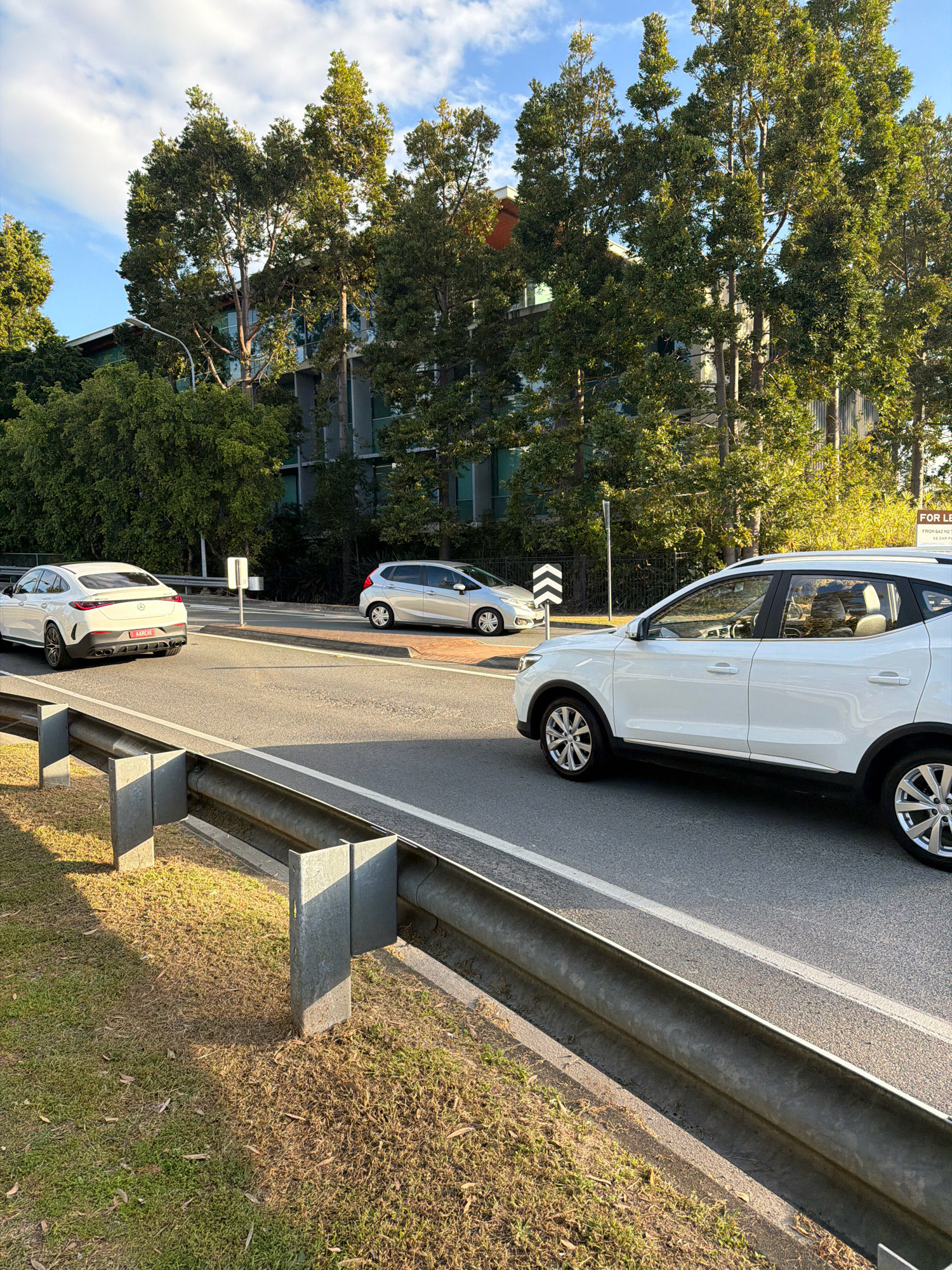 This image depicts a street scene in Broadbeach Waters, Australia. The foreground shows a metal guardrail bordering a grassy verge. Beyond the guardrail, a paved road with white lane markings is visible. Two white cars are present: a sleek sedan on the left, facing away from the camera, and an SUV on the right, also facing away. In the background, tall trees with dense green foliage line the street, partially obscuring a multi-story building with a modern architectural style featuring glass windows. The sky is blue with scattered clouds. A yellow "FOR LEASE" sign is partially visible in the far background on the right. The lighting suggests it is daytime, likely late afternoon due to the long shadows cast by the guardrail posts.