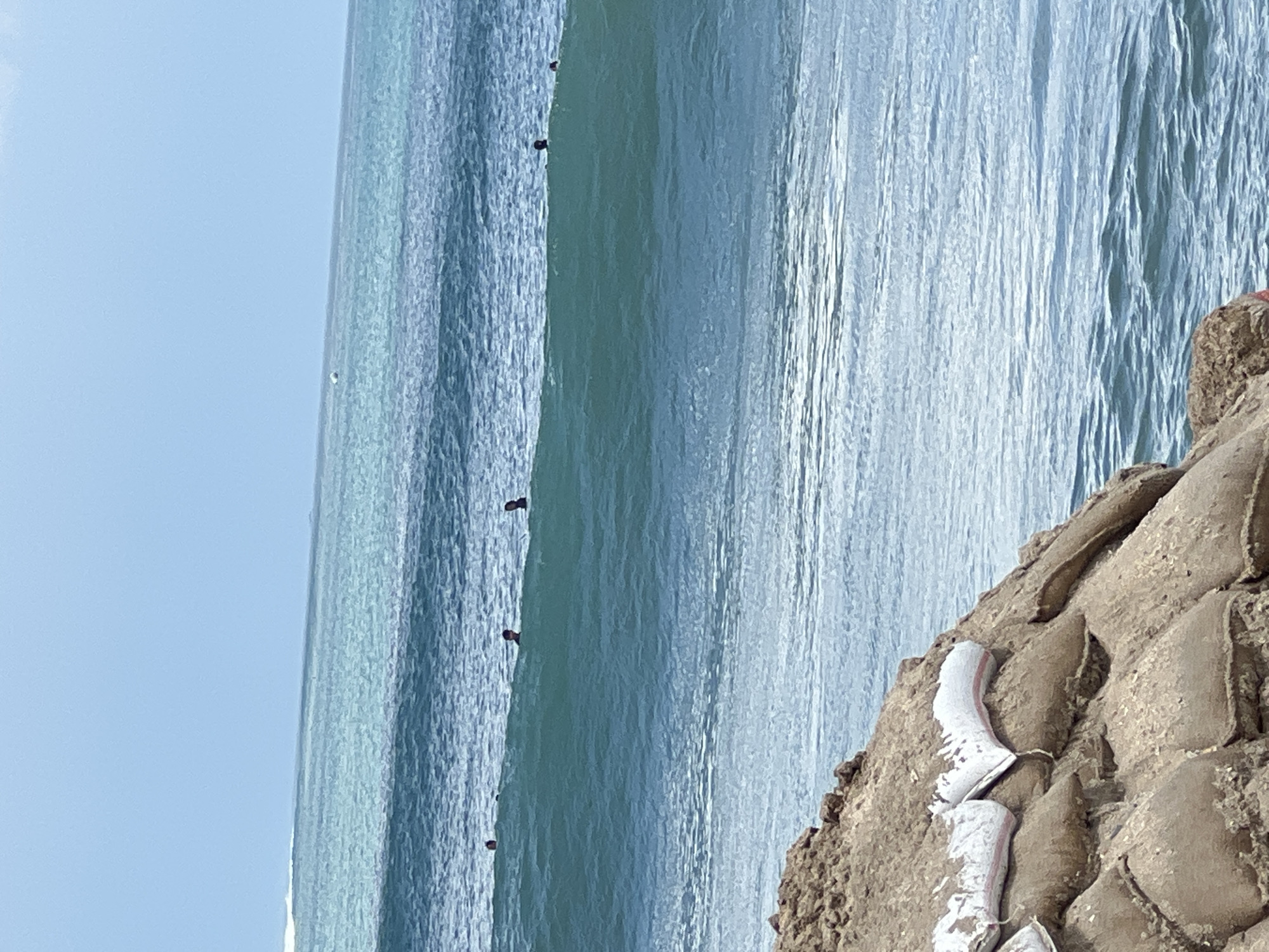 The image shows a view of the ocean with several people swimming or surfing in the water. In the foreground, there is a pile of sandbags, likely used as a barrier against the water. The sky is a light blue, and the water is a deep blue-green with several waves visible. The scene appears to be a beach or coastline. The location is Kuta, Indonesia.