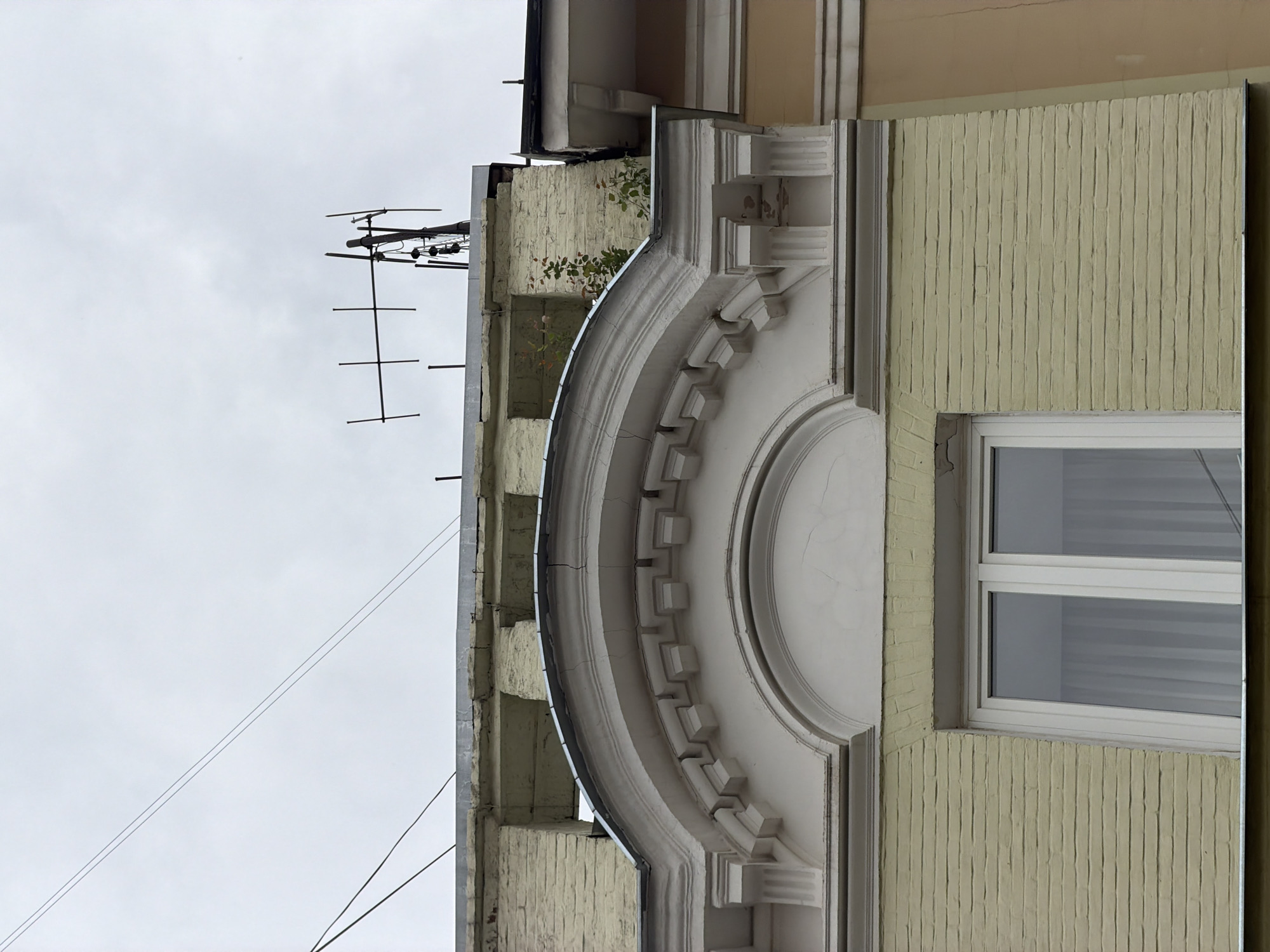 The image displays a detailed view of a building facade in Moscow, Russia, captured from a low angle against an overcast sky. The primary subject is a segment of an old building, featuring a prominent, elaborate white architectural decoration. This decoration consists of a large, semicircular arch-like molding with a central circular medallion and a band of regularly spaced square projections, reminiscent of dentils. The white stucco or stone surface of these decorative elements shows minor cracks and weathering.
The building's main wall is painted a light yellowish-beige color. In some sections, particularly around the window, the wall exhibits a distinct vertical ribbed texture, possibly indicating a specific type of rendered facade or painted brickwork. A modern white PVC-framed window with two panes is visible below the large architectural arch, reflecting interior details like horizontal blinds or stripes.
Above the main decorative arch, the upper part of the building reveals older, possibly brick, sections with peeling paint or plaster. Green foliage, resembling ivy or a small bush, grows on a ledge or balcony area. Against the light gray, cloudy sky, an old-fashioned directional antenna (likely for television or radio reception) is mounted on a structural element on the left side of the building. Several thin utility wires also stretch across the upper left portion of the sky. The overall scene is static, capturing architectural details and the urban environment on an overcast day. There are no people or visible text in the image.