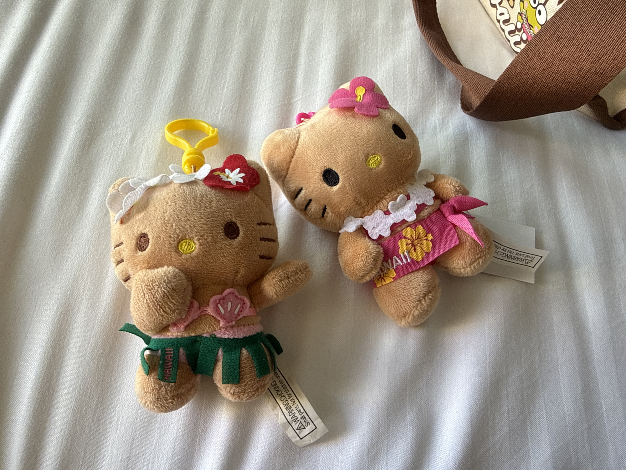 Two plush Hello Kitty dolls are lying on a white, slightly wrinkled surface. One doll wears a green hula skirt, a seashell necklace, and a flower in its hair, with a yellow plastic hook attached to its head. The other doll is dressed in a pink dress with floral accents and a matching flower in its hair. Both dolls have brown fur and are lying next to a brown bag strap. A tag with a warning label is visible near the doll with the hula skirt. The location is unknown.