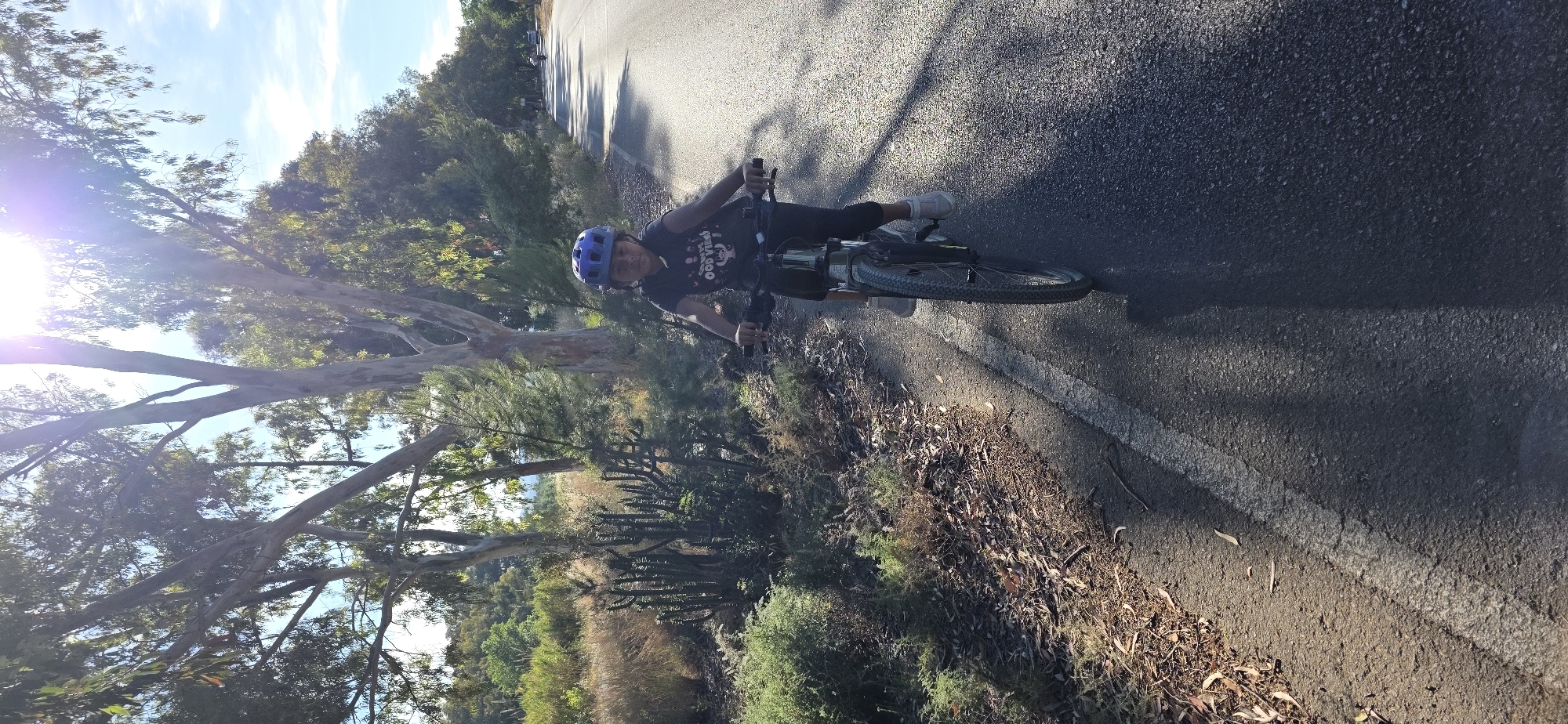 The image shows a person riding a bicycle on an asphalt road. The person is wearing a blue helmet, a dark-colored t-shirt, and dark pants. They are wearing white shoes. The bicycle is a mountain bike. The road has a white line marking the edge. To the side of the road is vegetation consisting of trees, bushes, and dry leaves. The sky is visible with some clouds and a lens flare from the sun.
Without specific identifying features such as landmarks, the precise location in Canet d'en Berenguer cannot be definitively determined. However, the scene suggests a semi-rural or park area with a paved road suitable for cycling.